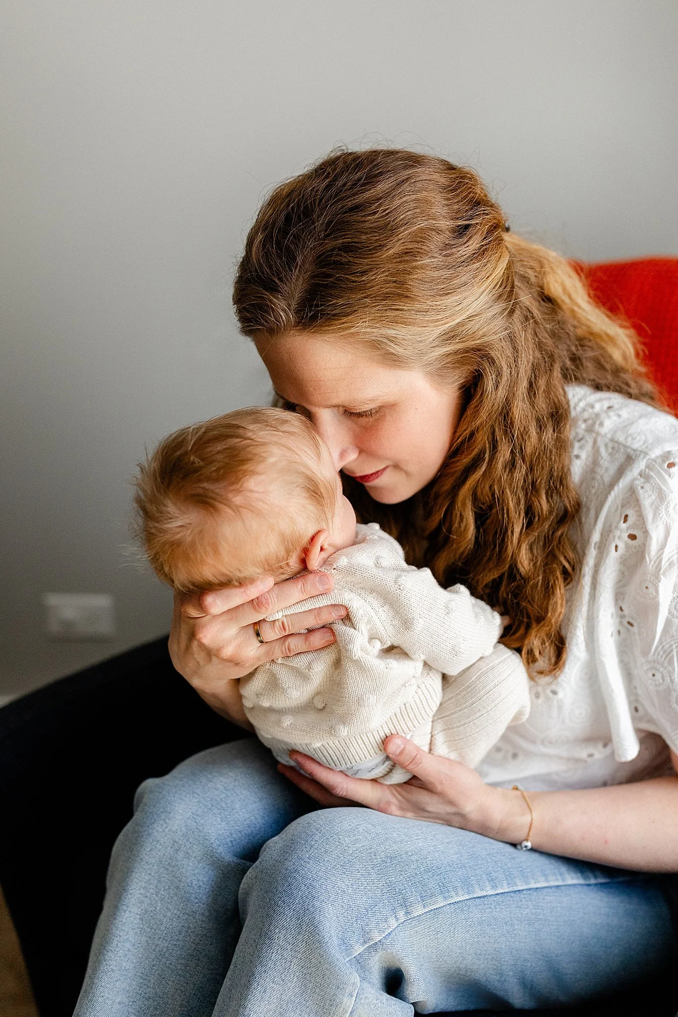 A woman with long, wavy brown hair is holding and kissing a young child with blonde hair. They are sitting together indoors, sharing an intimate moment, with the woman wearing a white embroidered blouse and blue jeans with newborn photographers.