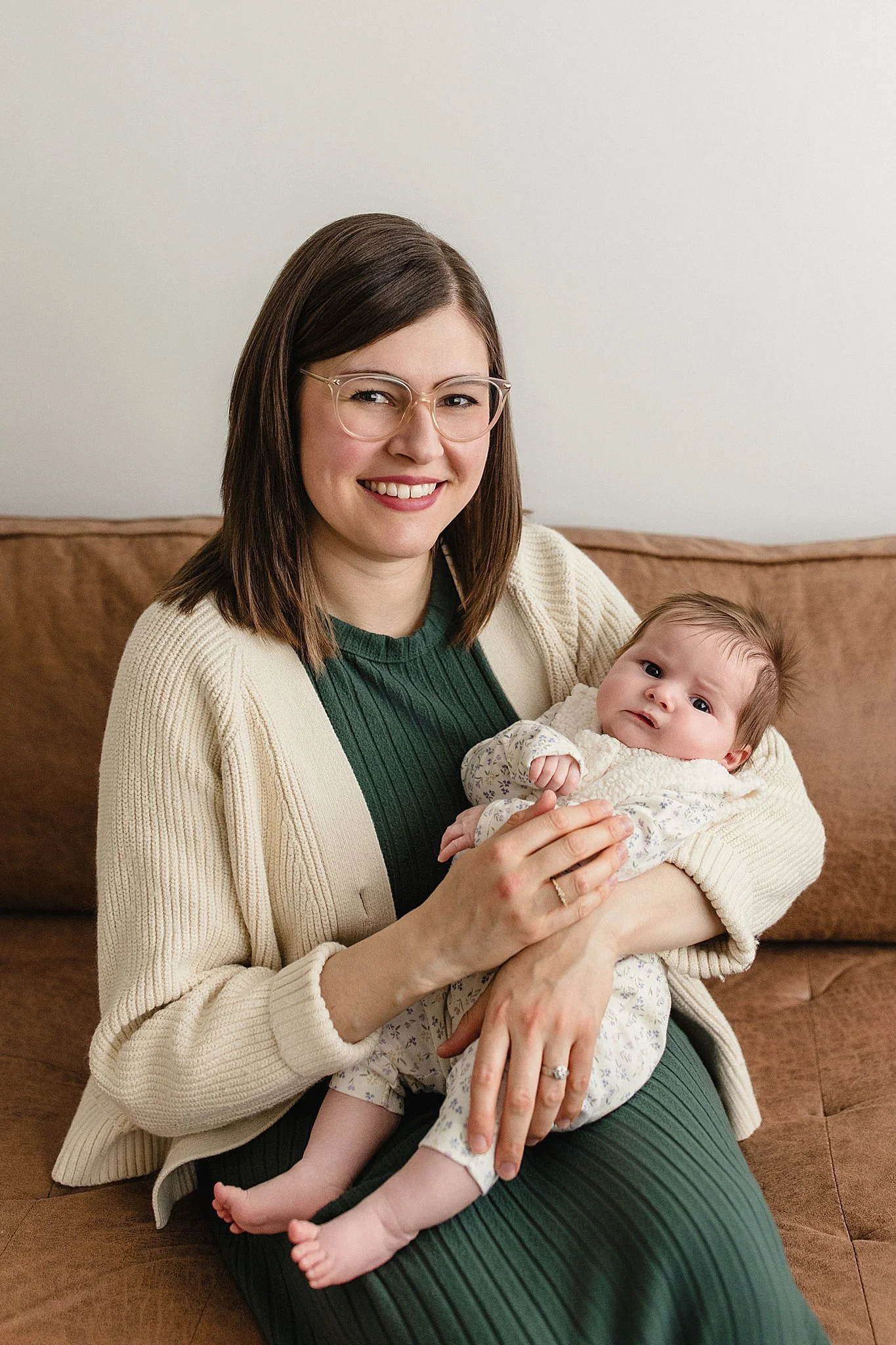 A woman with shoulder-length brown hair, glasses, and a beige cardigan holding a baby girl on a brown couch, both looking at the camera during a studio family photography session in downtown Naperville.