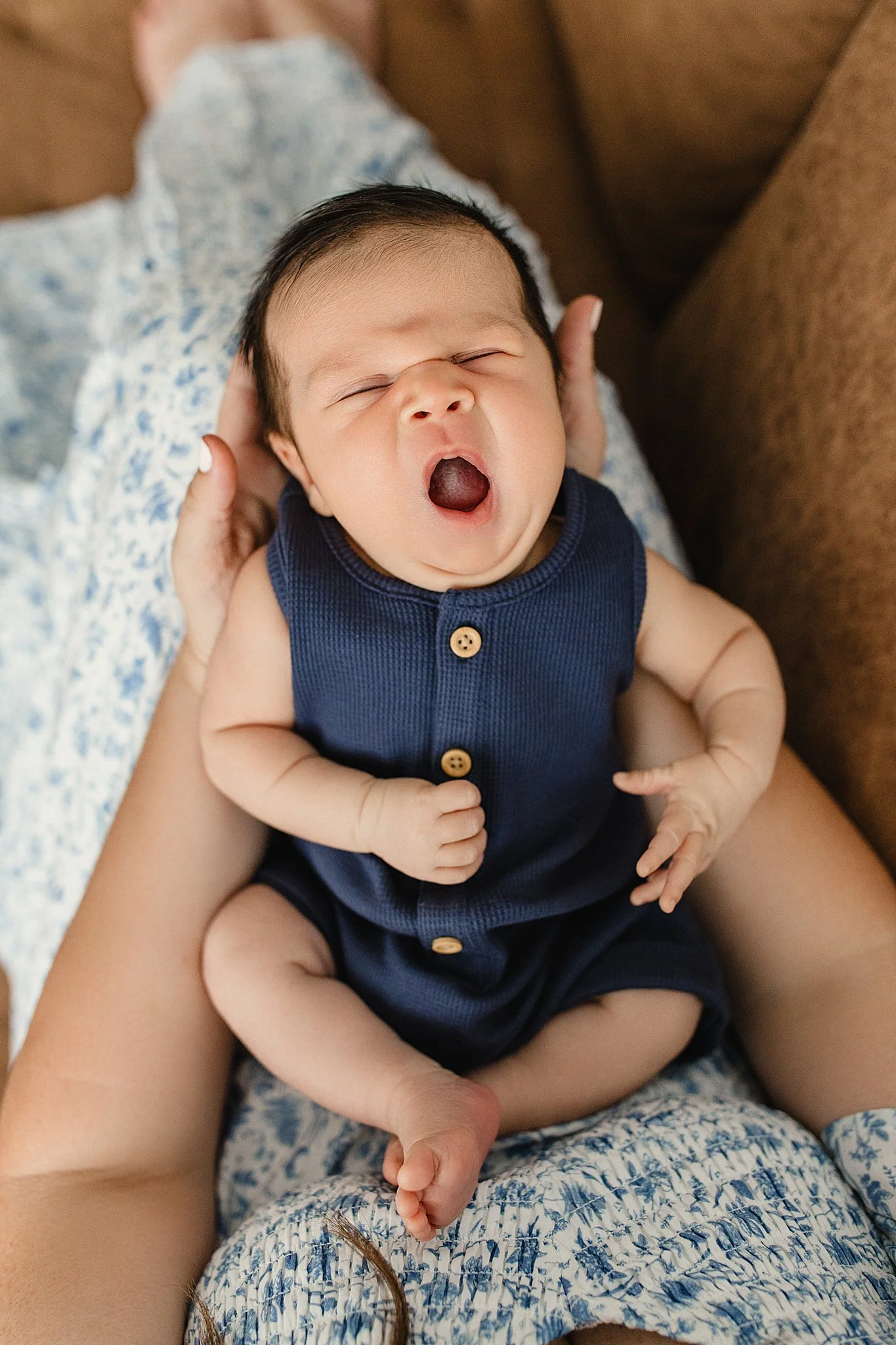 A baby with dark hair yawning, being held by someone. The baby is wearing a navy blue sleeveless outfit with wooden buttons, and they are sitting on the person's lap, which is covered with a blue and white patterned fabric with a newborn photographer