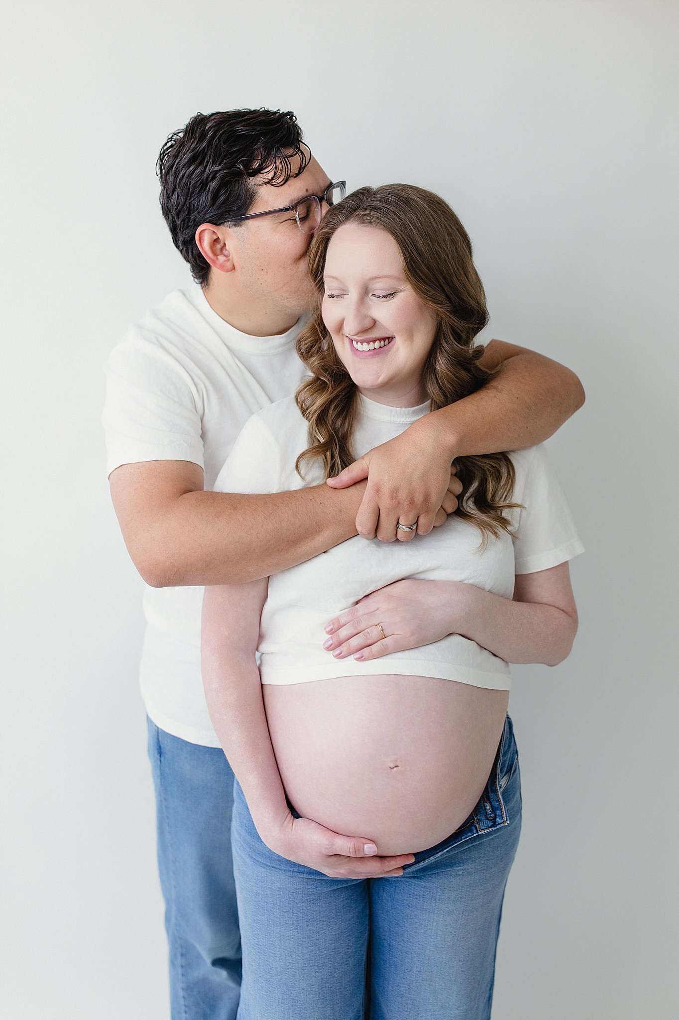 A pregnant woman with long brown hair and a man with short dark hair and glasses embrace in front of a plain white wall. The man kisses the woman's forehead while she smiles and rests her hands on her pregnant belly during Naperville maternity photos