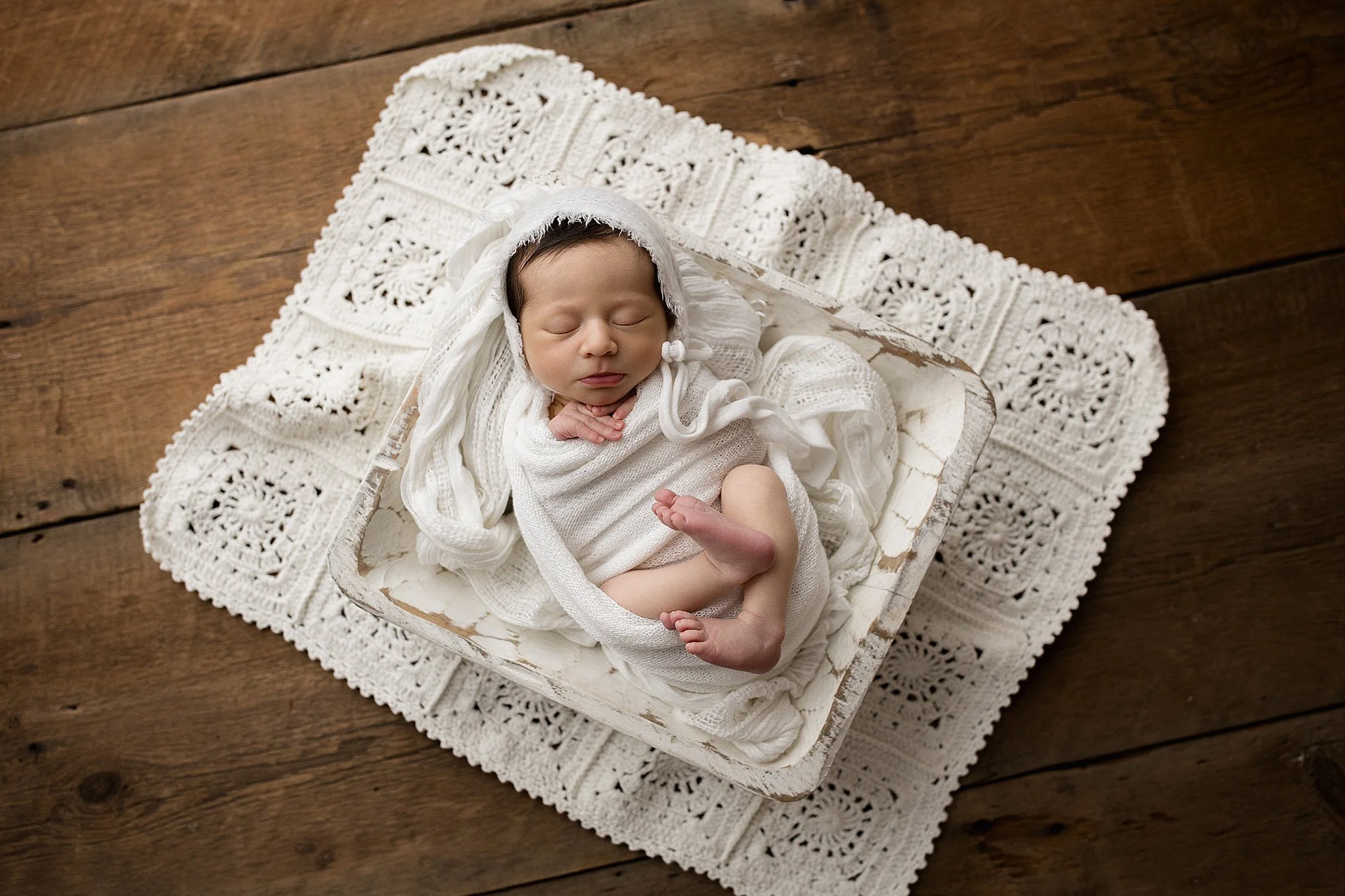 A newborn baby sleeping on a white lace blanket and crochet mat, wrapped in white fabric with a bonnet, on a wooden surface.