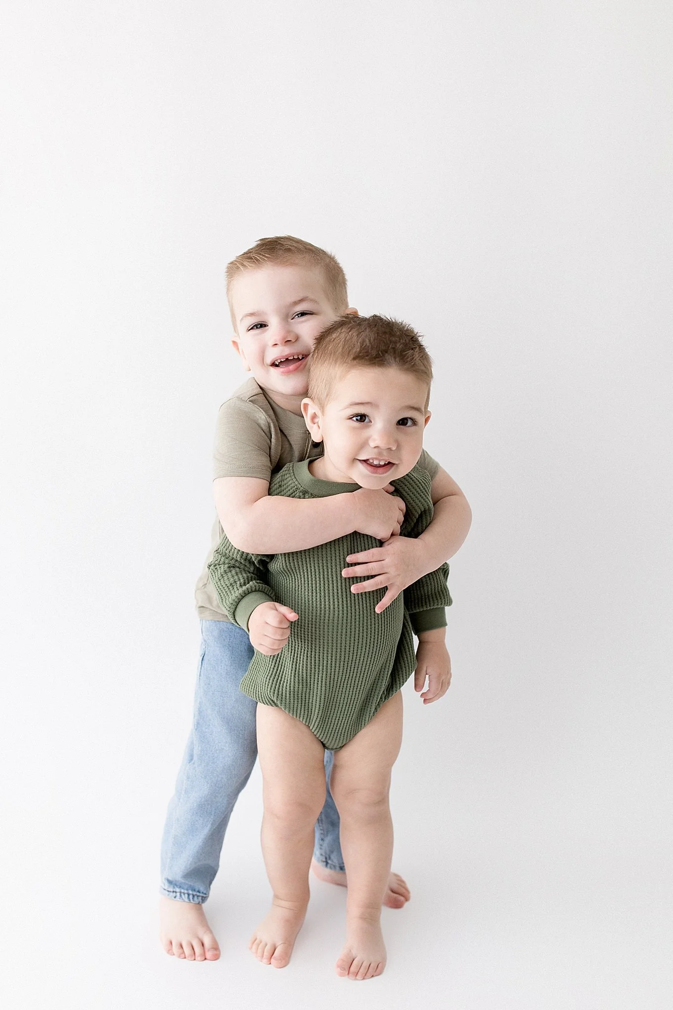 Two young boys, one older and one younger, smiling and hugging each other against a white background kids studio photography.