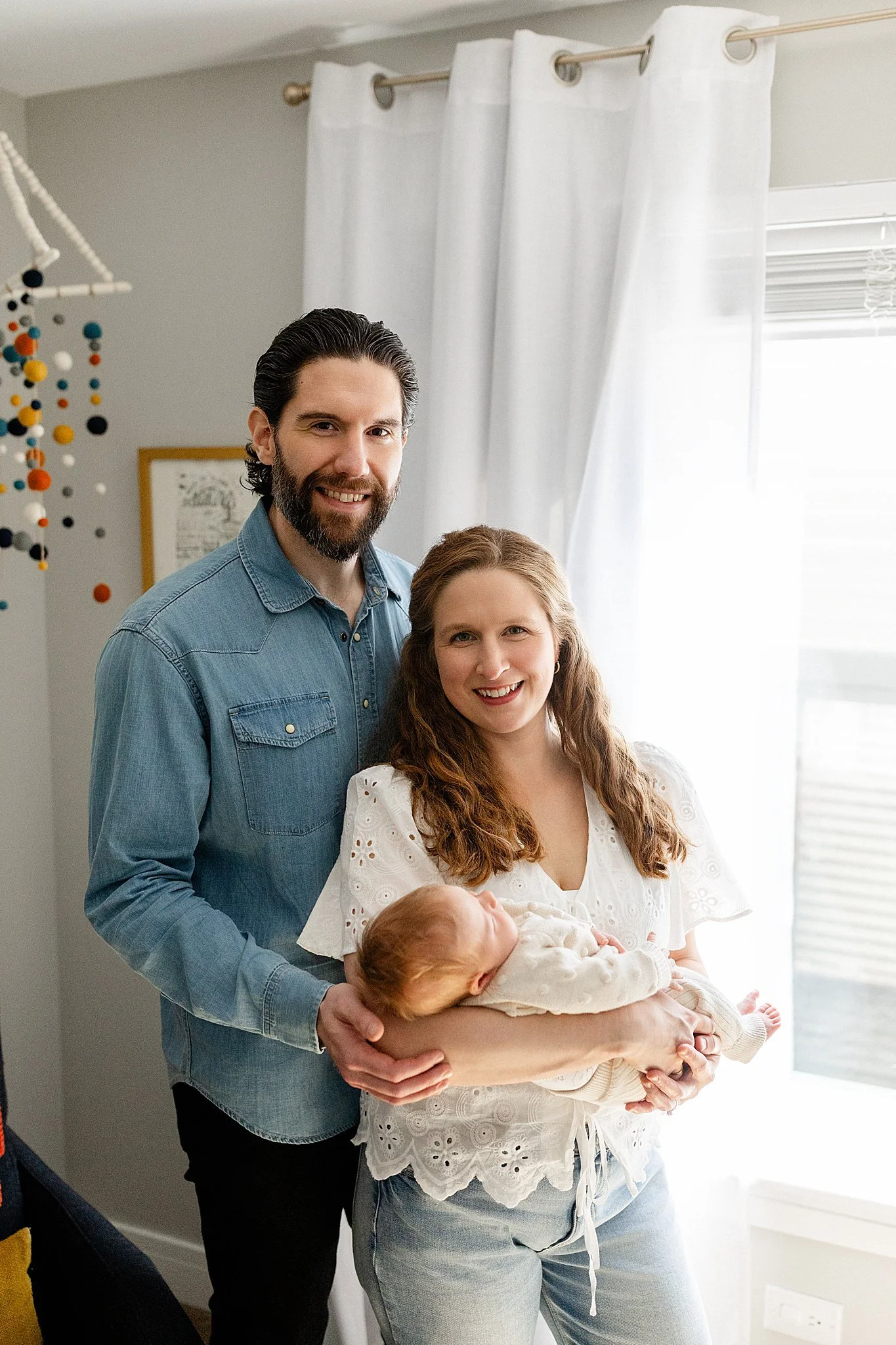 A smiling man with dark hair and beard, wearing a denim shirt, stands beside a woman with long curly red hair who is holding a baby wrapped in a white blanket for lifestyle newborn photos in Hinsdale, IL.