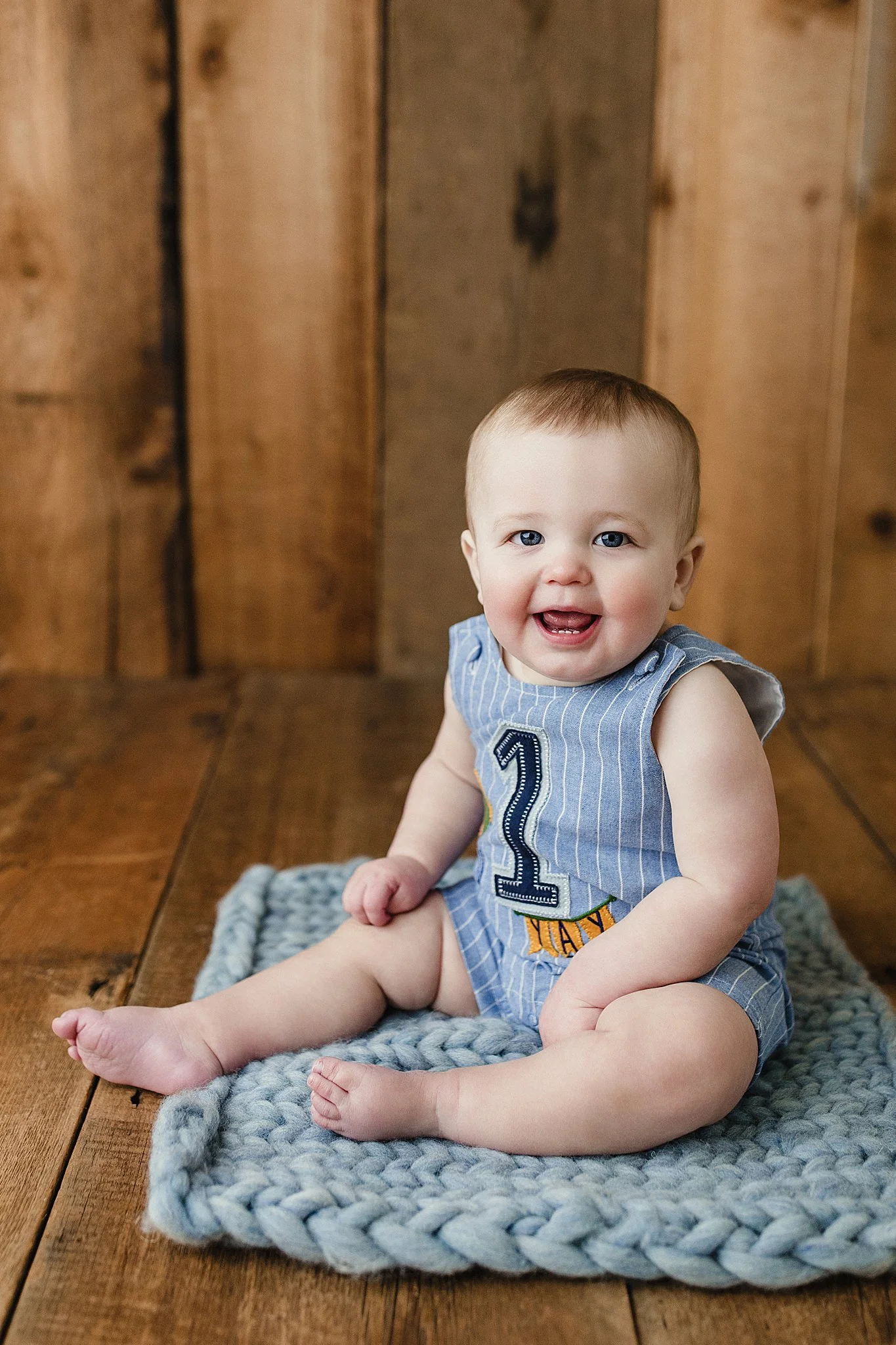 A smiling baby sitting on a gray knitted blanket on a wooden floor, wearing a blue striped sleeveless outfit with a number 1 on it and orange letters for a first birthday photoshoot in Naperville, IL.