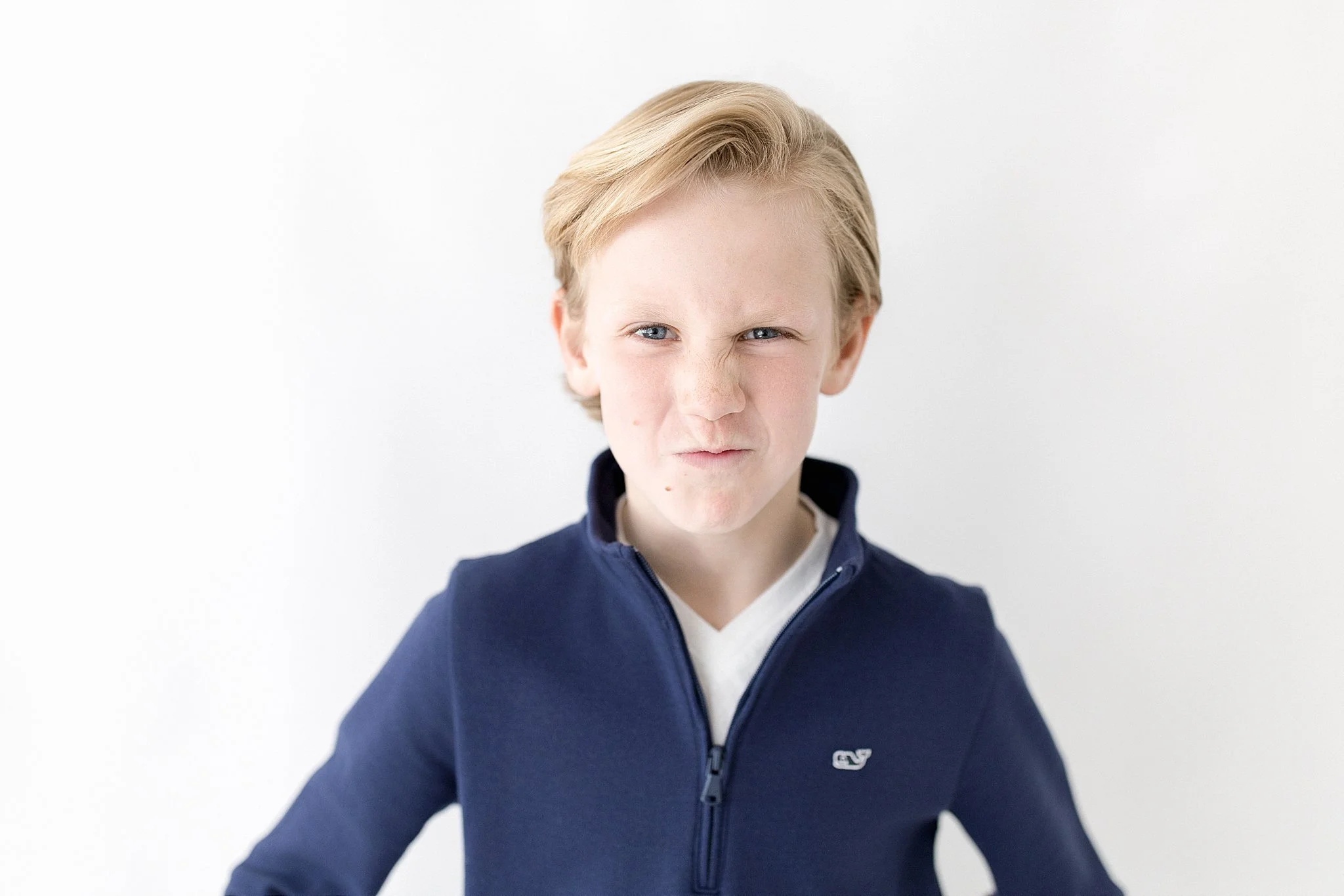 A boy with blonde hair and blue eyes making a scrunched-up facial expression, wearing a navy blue zip-up jacket and a white shirt underneath, standing against a white background with the best photographer in Naperville, IL.