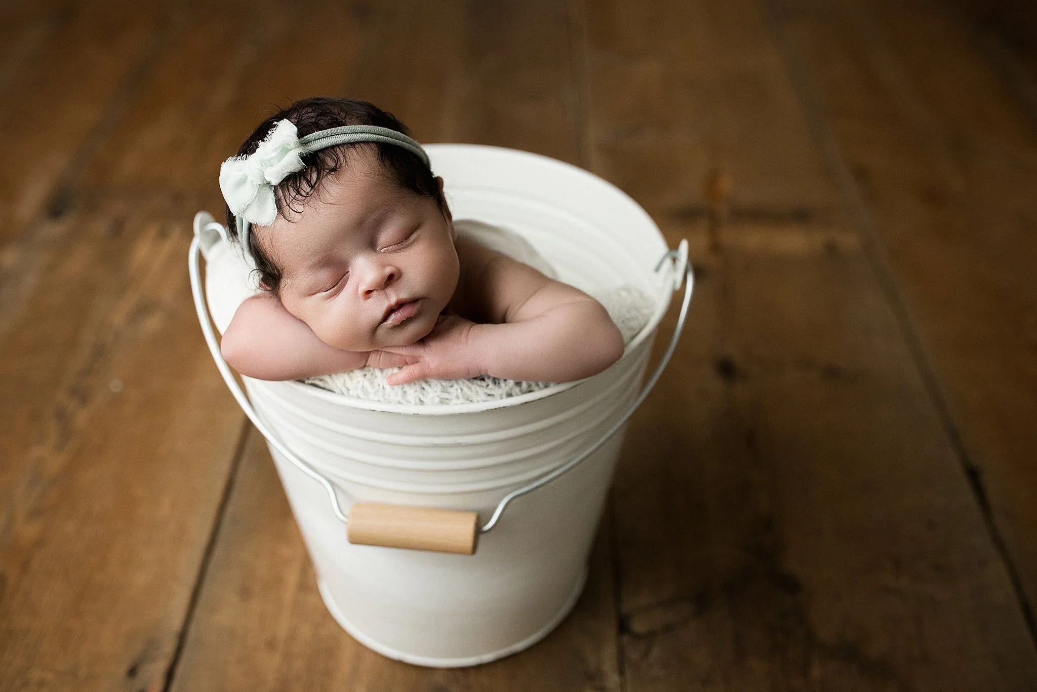 A sleeping newborn baby with dark curly hair, wearing a gray headband with a white flower, is resting with arms folded on a white blanket inside a white bucket on a wooden floor during posed newborn photography near me.