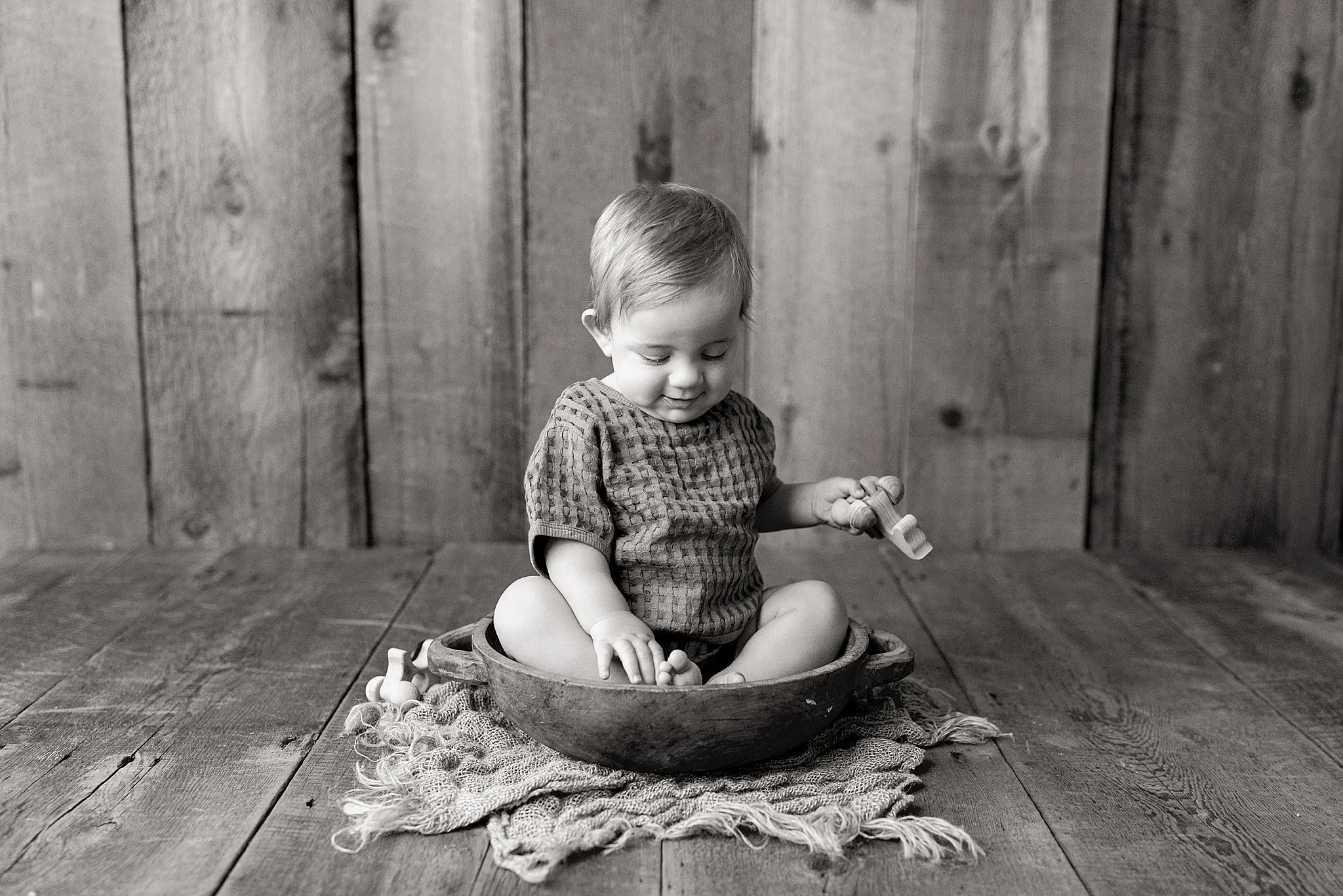 A young child sitting on a wooden floor, playing with toys in a round wooden bowl, with a rustic wooden wall in the background for a baby photoshoot in Naperville, IL.