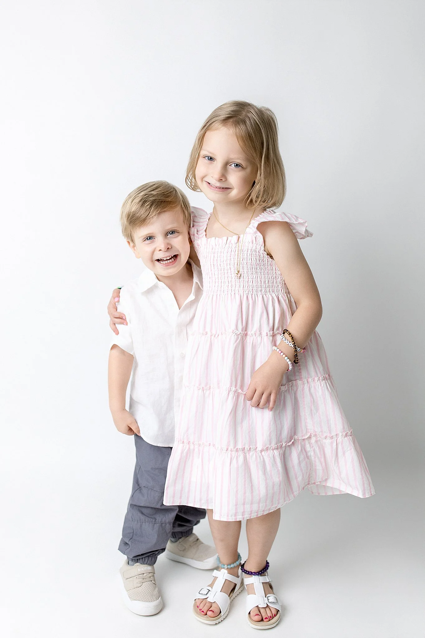 Two young children, a girl and a boy, standing close together against a plain white background. The girl is wearing a pink and white striped dress with ruffled sleeves and white sandals, while the boy is dressed in a white shirt, gray pants, and beig