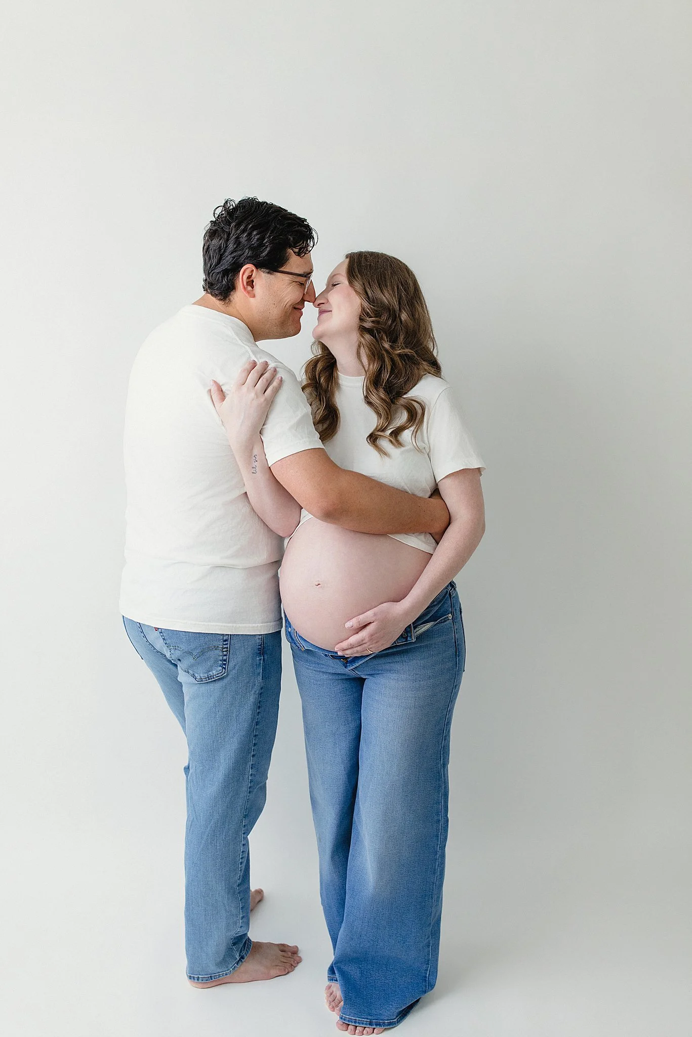 A pregnant woman and a man are standing close together with their noses touching, smiling, and embracing against a plain white wall. They are wearing casual white t-shirts and jeans, both barefoot with maternity photographers near me.