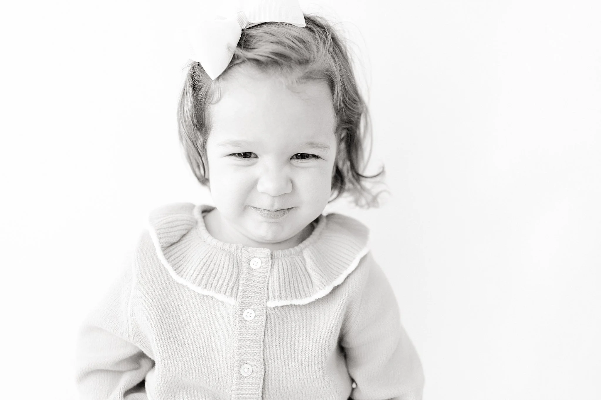 A young girl with a bow in her hair, wearing a sweater, smiling gently while looking at the camera with a photographer who does personality minis.