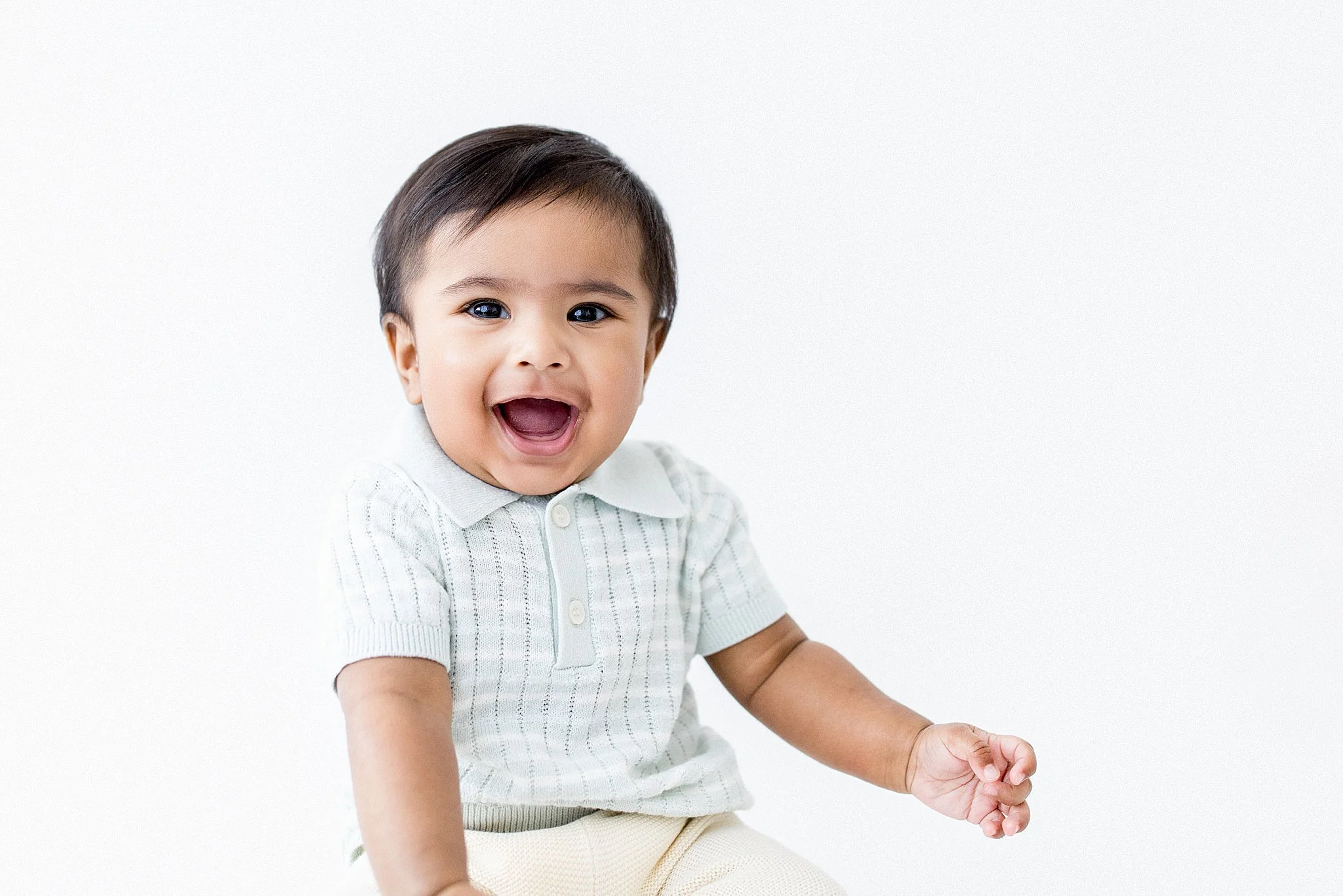 Smiling young boy with dark hair and big eyes, wearing a white collared shirt, sitting against a plain white background during baby photos in Naperville.