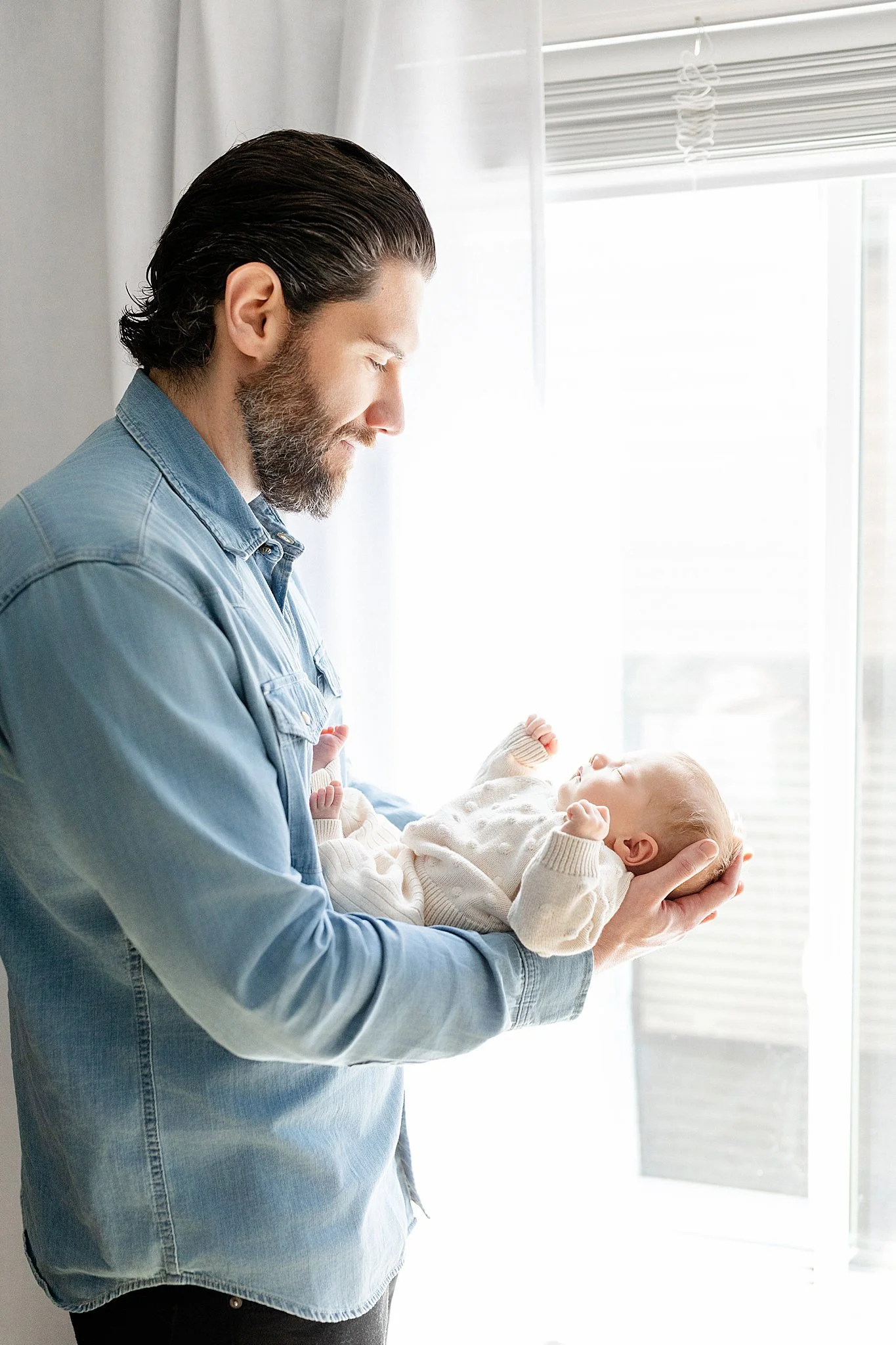 A man with dark hair and a beard holding a baby, both with closed eyes, in front of a window with white blinds and curtains, during daytime with a photographer near me.
