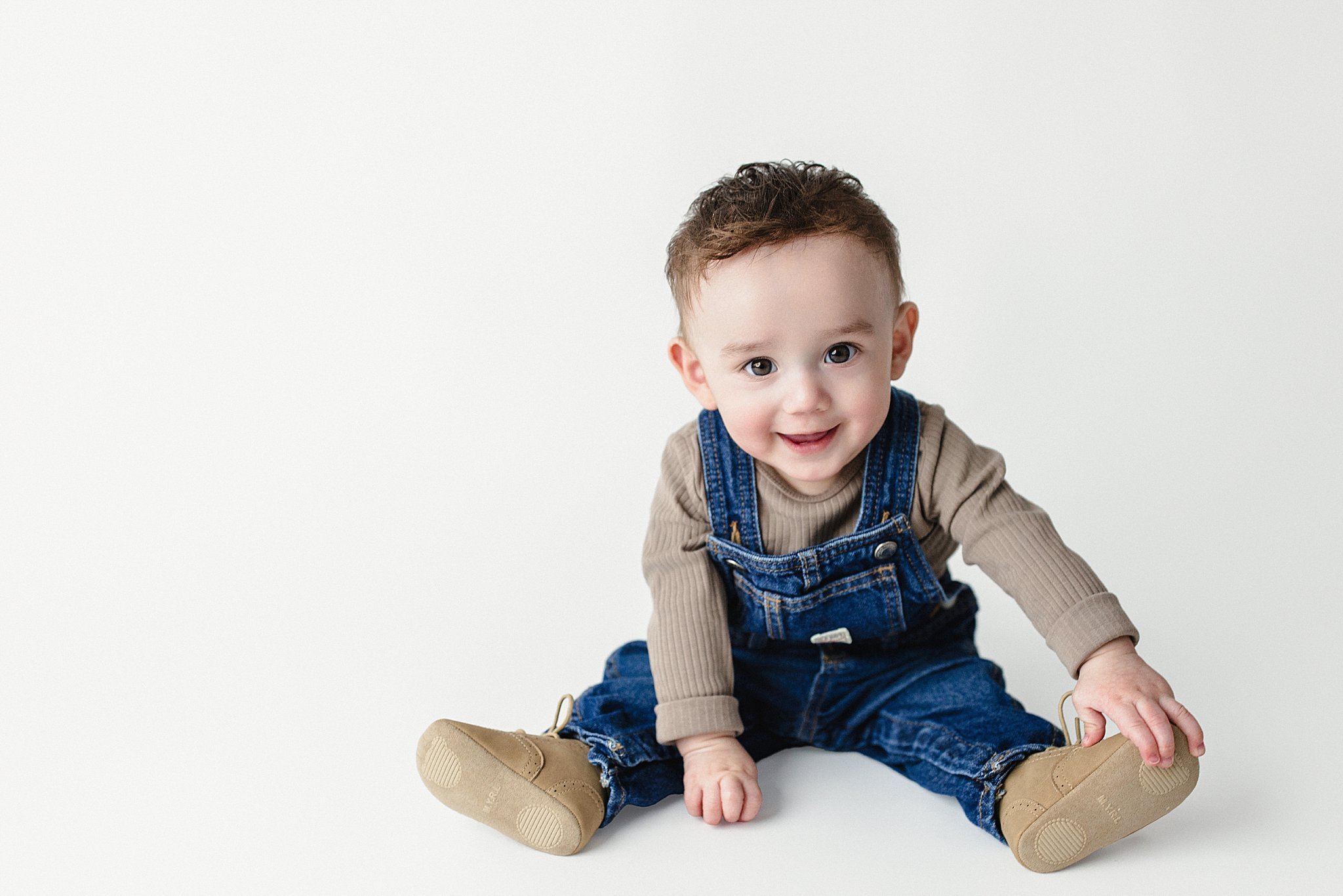 A smiling toddler sitting on the floor with legs spread apart, wearing beige shoes, denim overalls, and a beige long-sleeve shirt, against a plain white background for baby portraits near me.