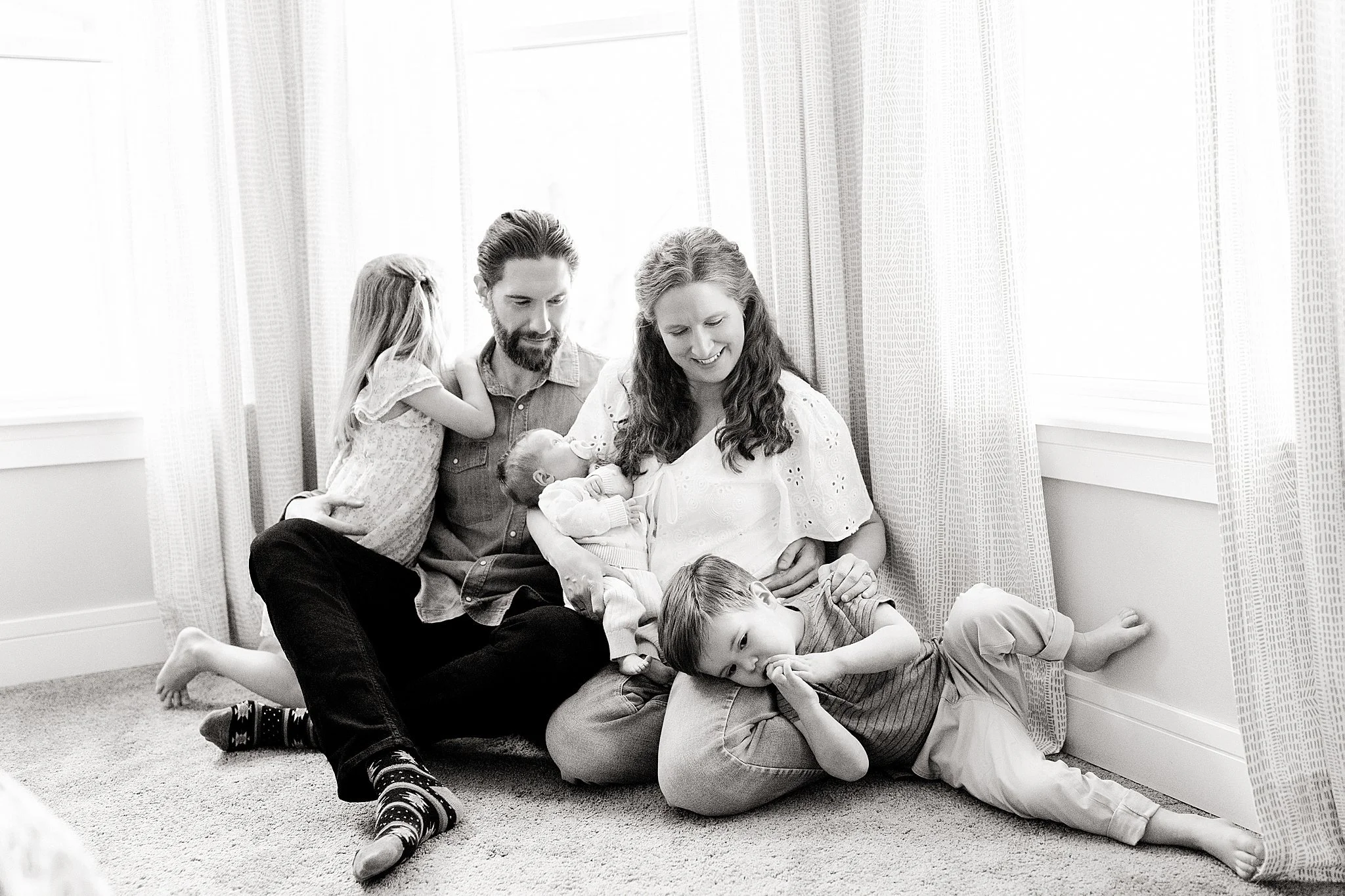 Black and white photo of a family sitting on the floor in a room with curtains. Two adults and four children are relaxing, with some of the children lying on top of the adults.