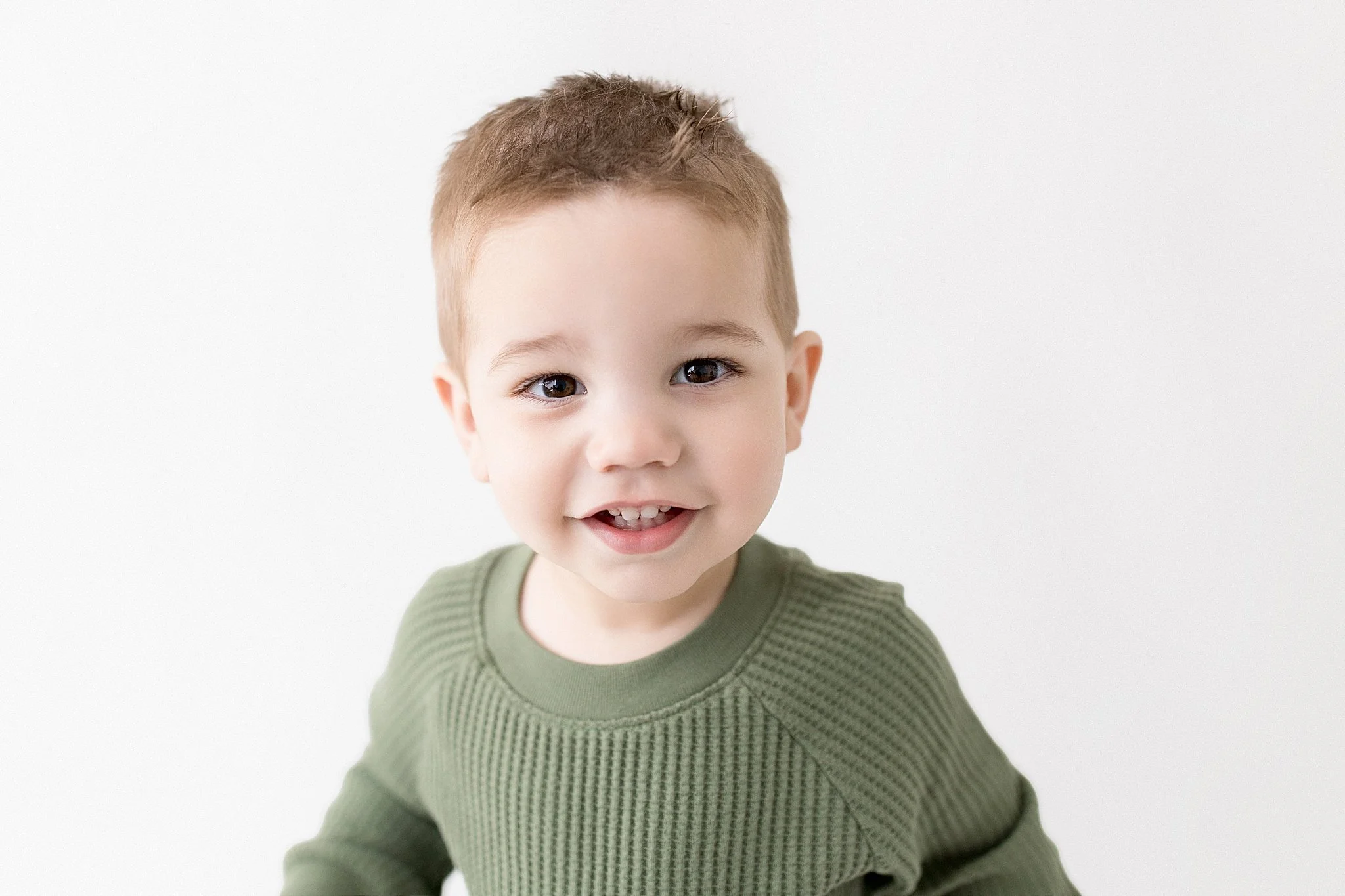 Smiling young boy with short light brown hair wearing a green sweater against a white background for baby photographers near me.