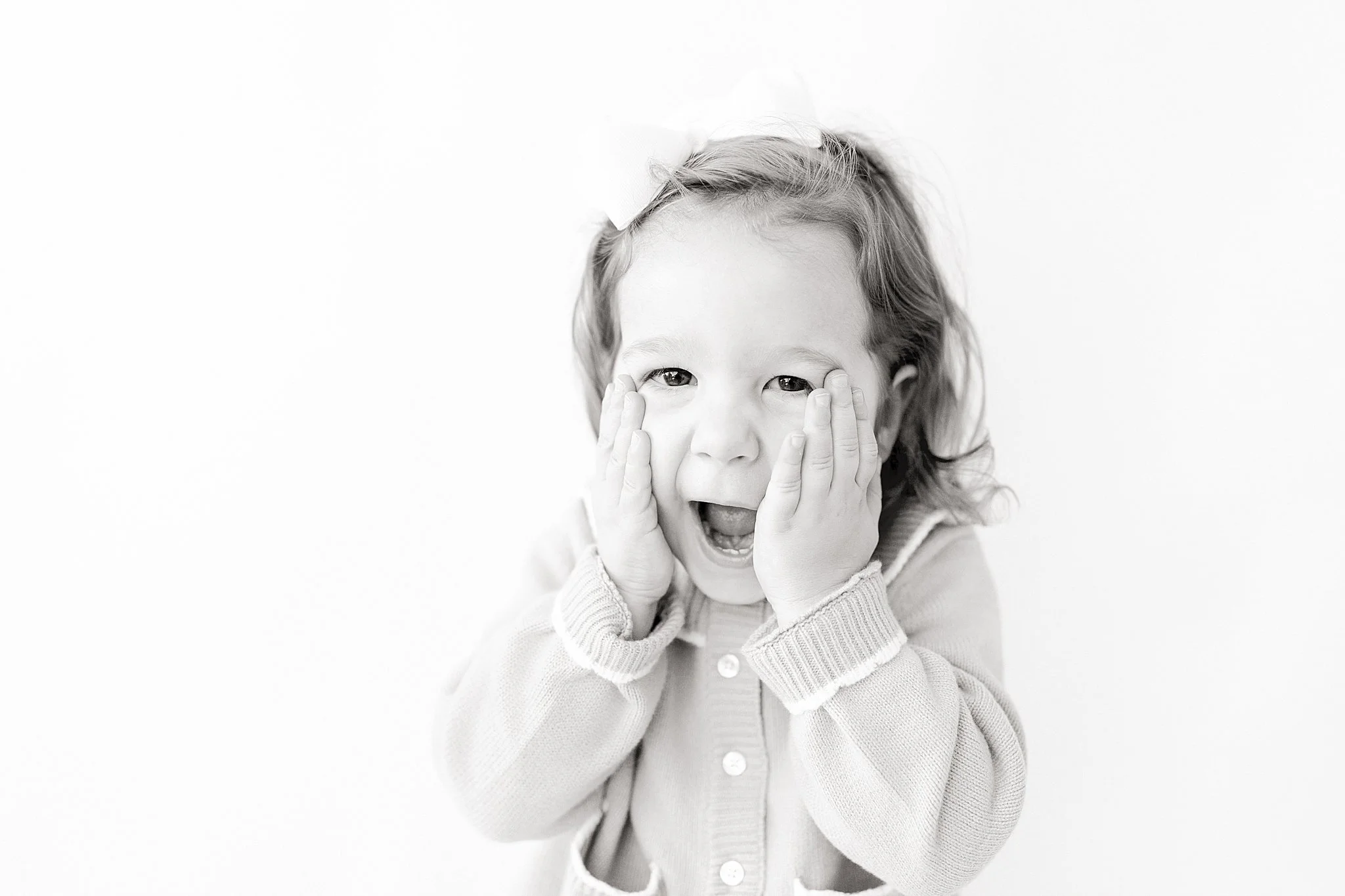 Black and white photo of a young girl with curly hair, wearing a sweater, holding her face with both hands, and showing an excited or surprised expression during a personality portrait fundraiser event with Ally and B Photography.