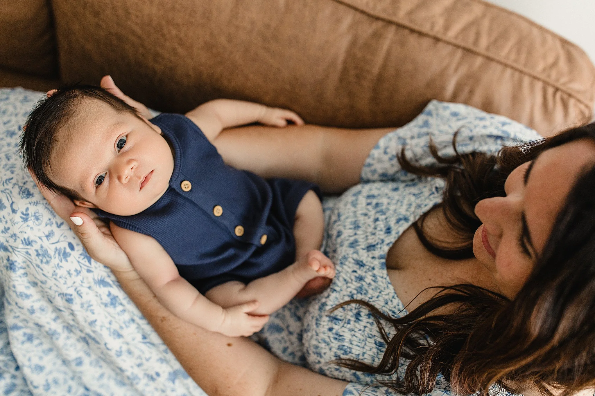 A baby lying on an adult's chest on a floral pillow, looking at the camera, with an adult woman lying next to him, partially visible with long brown hair with newborn photographers near me.