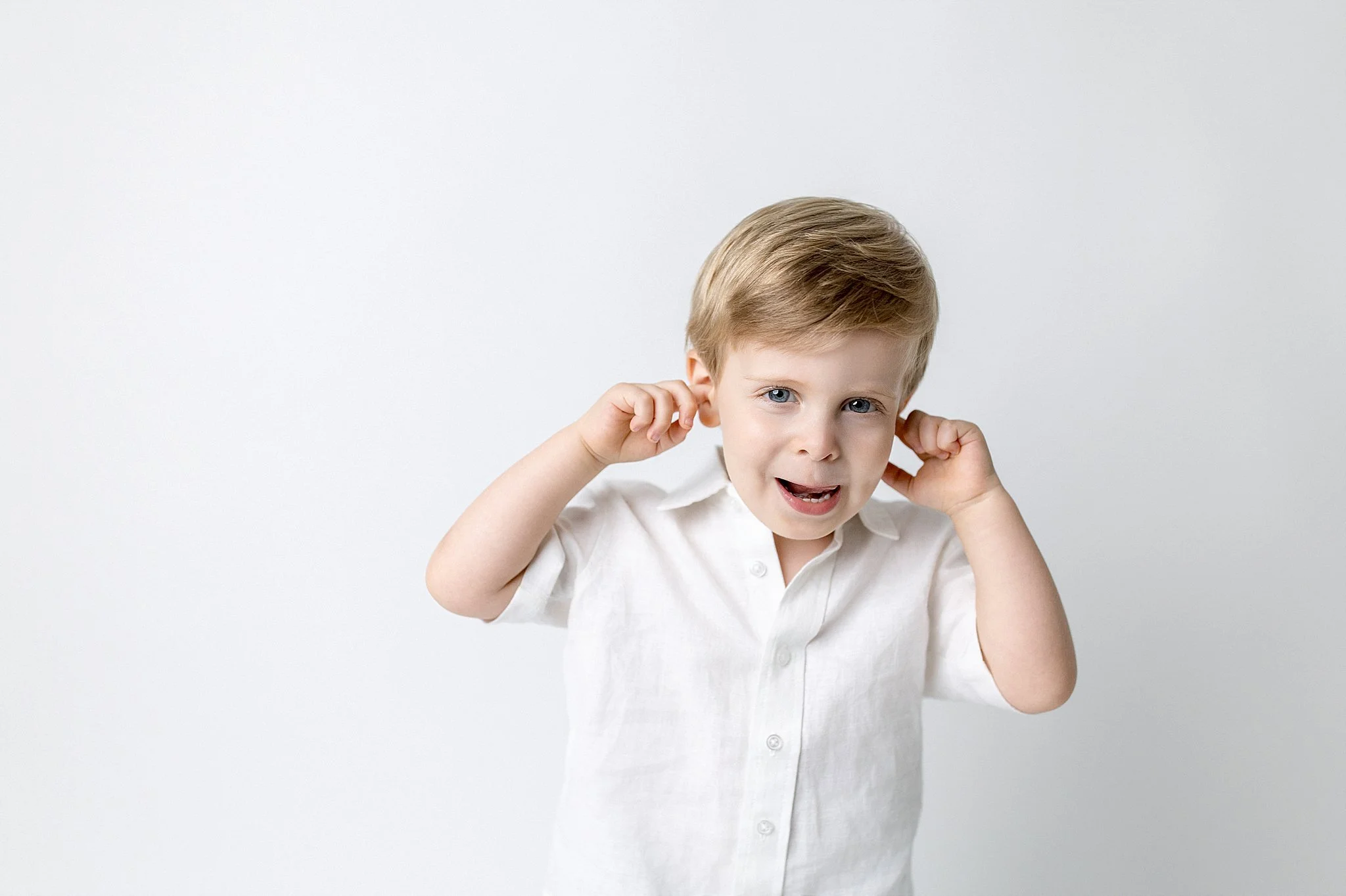 A young boy with blonde hair and blue eyes wearing a white shirt, covering his ears and looking at the camera, standing against a plain white background during a downtown Naperville photoshoot.