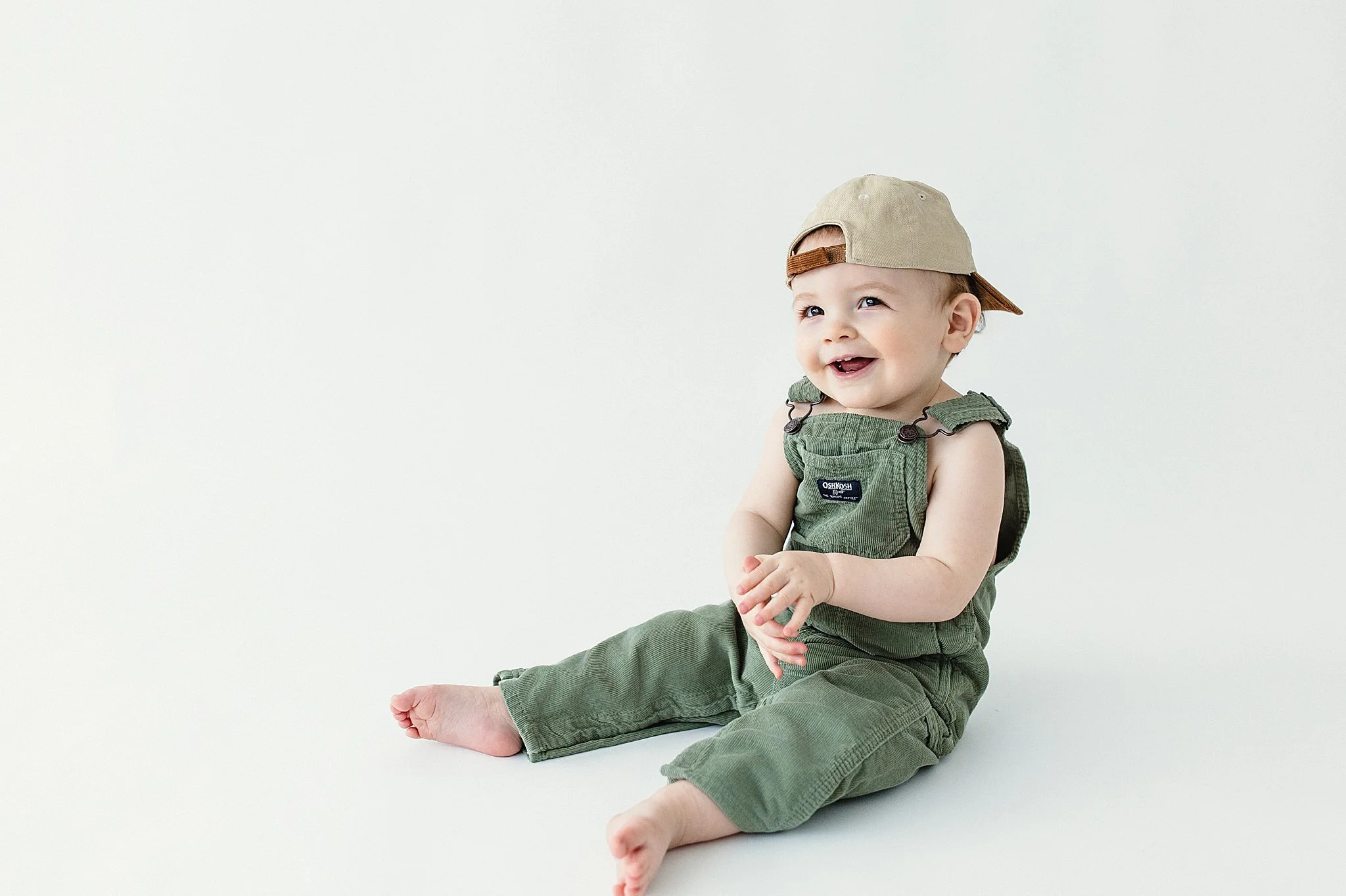 Smiling baby boy sitting on the floor, wearing green overalls and a tan baseball cap, against a plain white background during a milestone photoshoot in Naperville with Ally and B Photography.