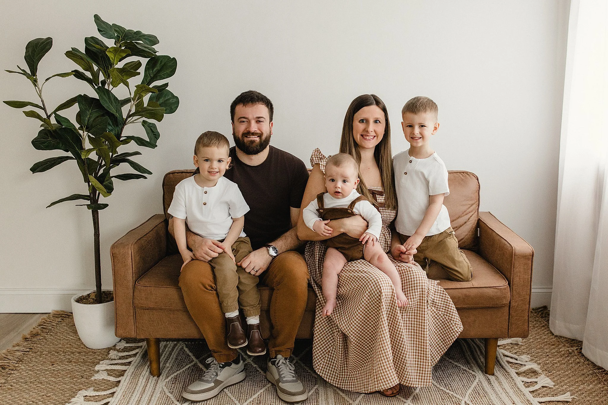A family of six sitting on a brown couch in a living room, smiling at the camera. The parents are in the middle, with three children, two boys and a baby girl, sitting around them during a lifestyle family photo session near me.