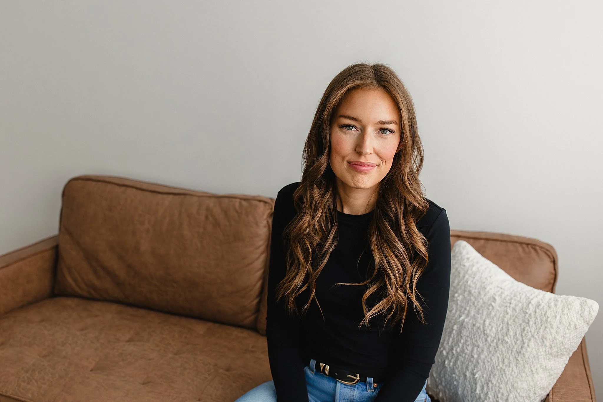 A young woman with long wavy brown hair and light skin, wearing a black top and blue jeans, sitting on a brown couch in a room with white walls for influencer brand photos.
