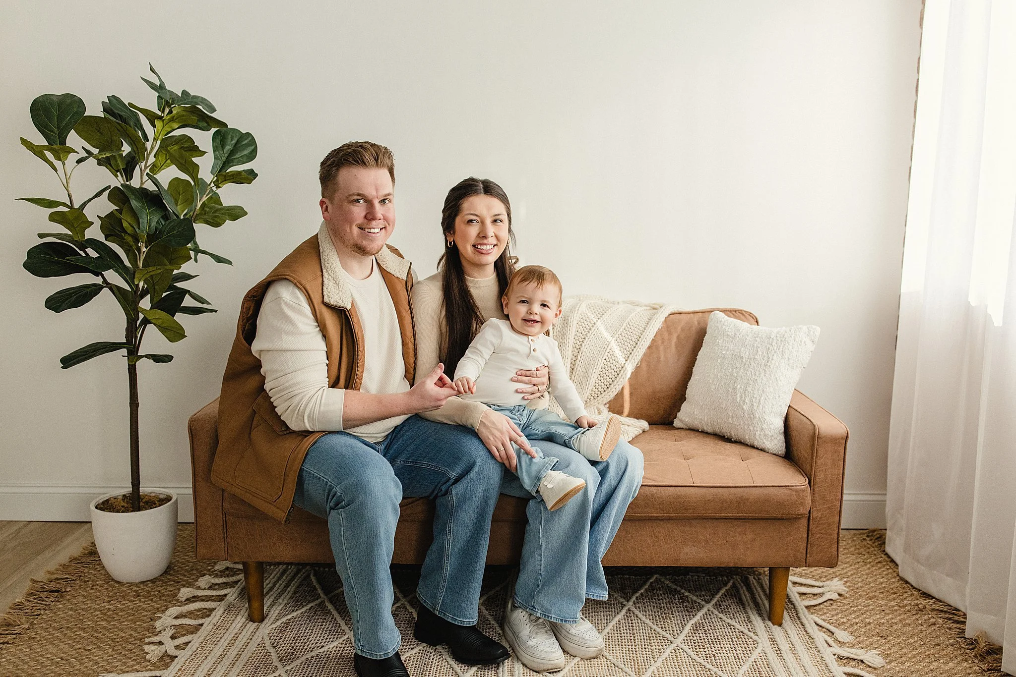 A happy family of three sitting on a beige sofa in a bright living room with a white wall, a potted plant, and white curtains. The family includes a man, woman, and a young child during lifestyle photos near me.