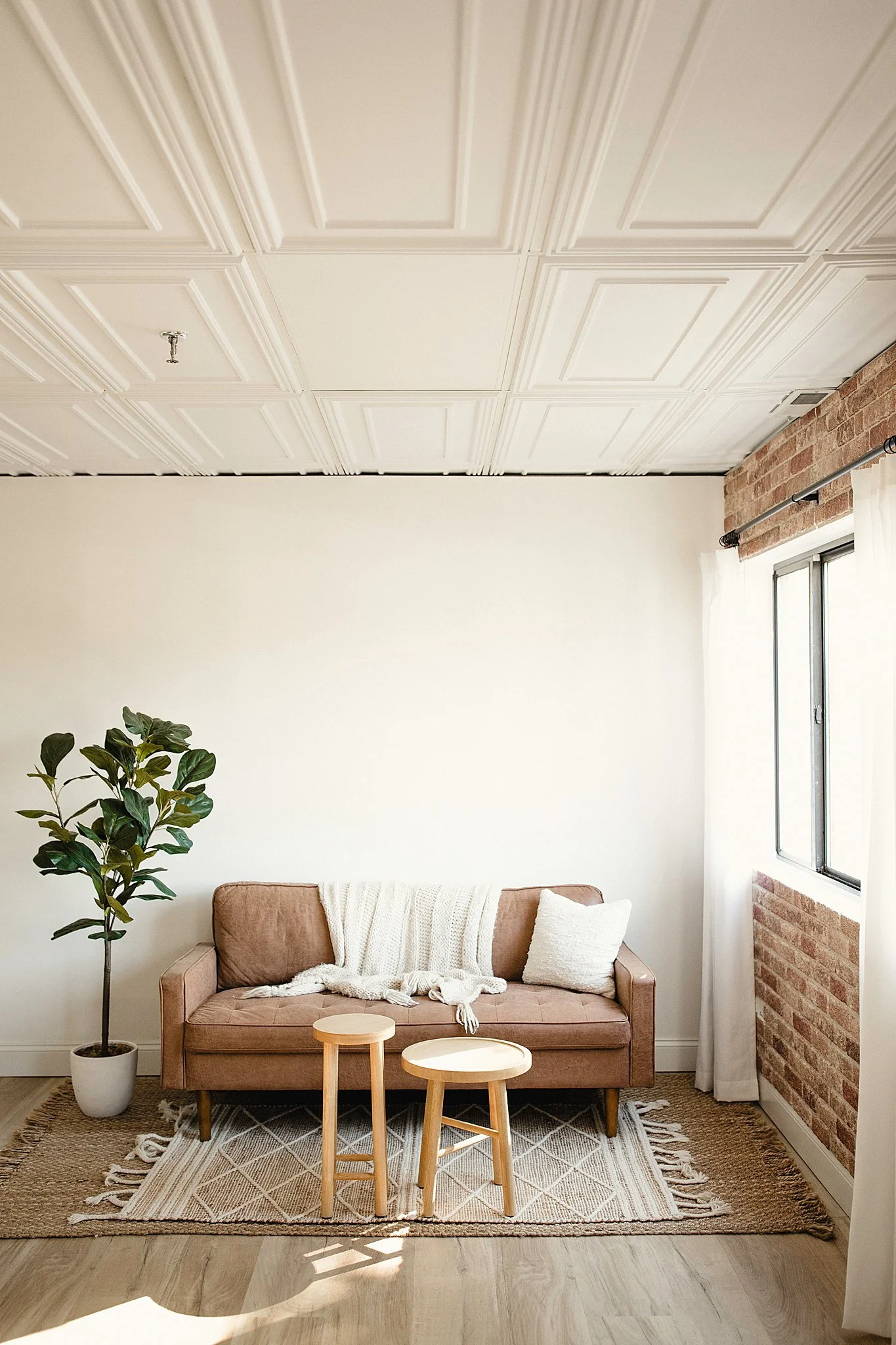 Living room with a light brown sofa, white cushion, and throw blanket, potted plant, two small wooden stools, patterned rug, brick wall, window with curtains, and a white ceiling with paneling at Ally and B Photography studio in Naperville, IL.