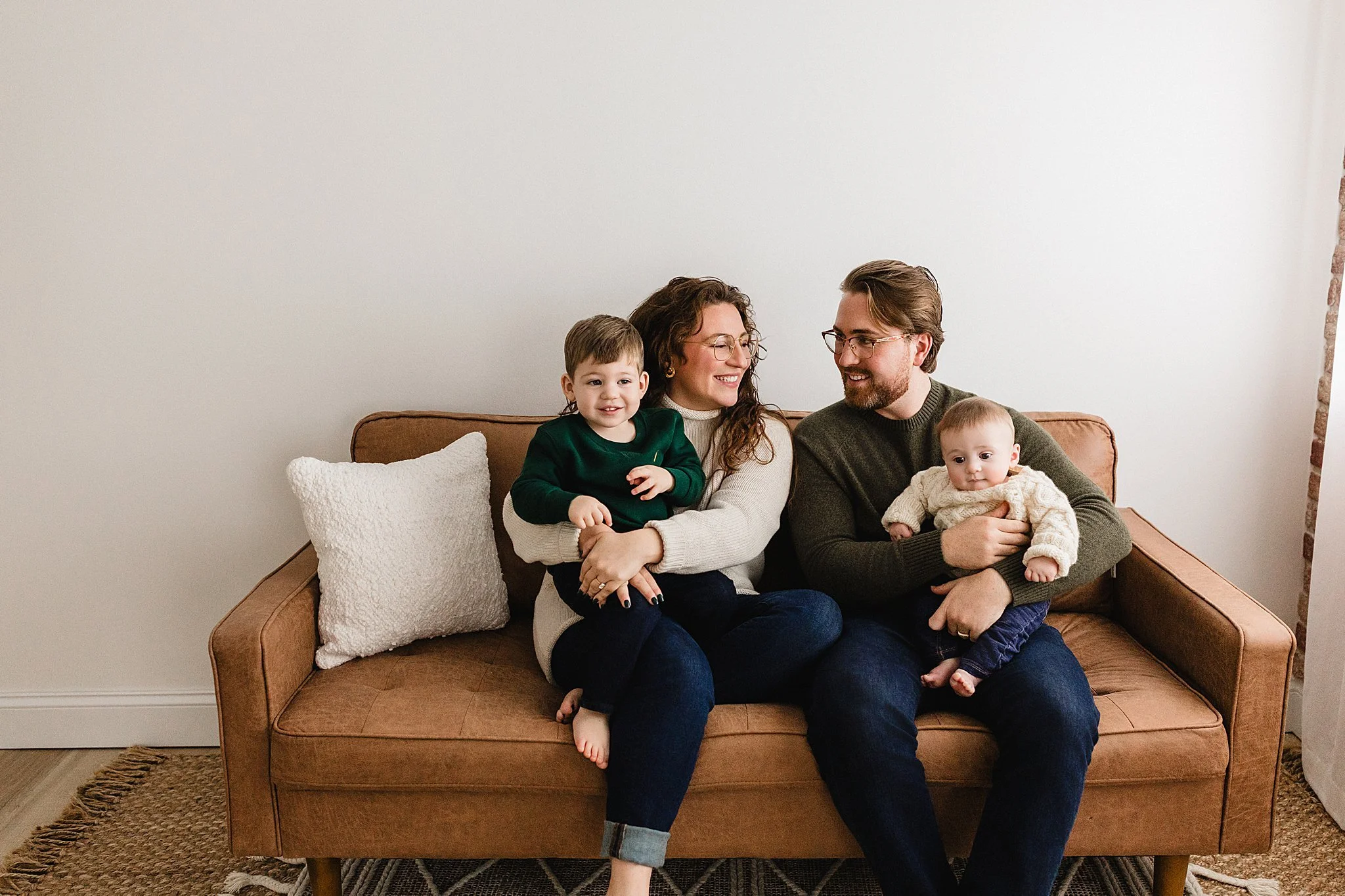A family of four sitting on a brown couch, smiling and enjoying each other's company in a living room during a studio photoshoot with Ally and B Photography in downtown Naperville, Illinois.