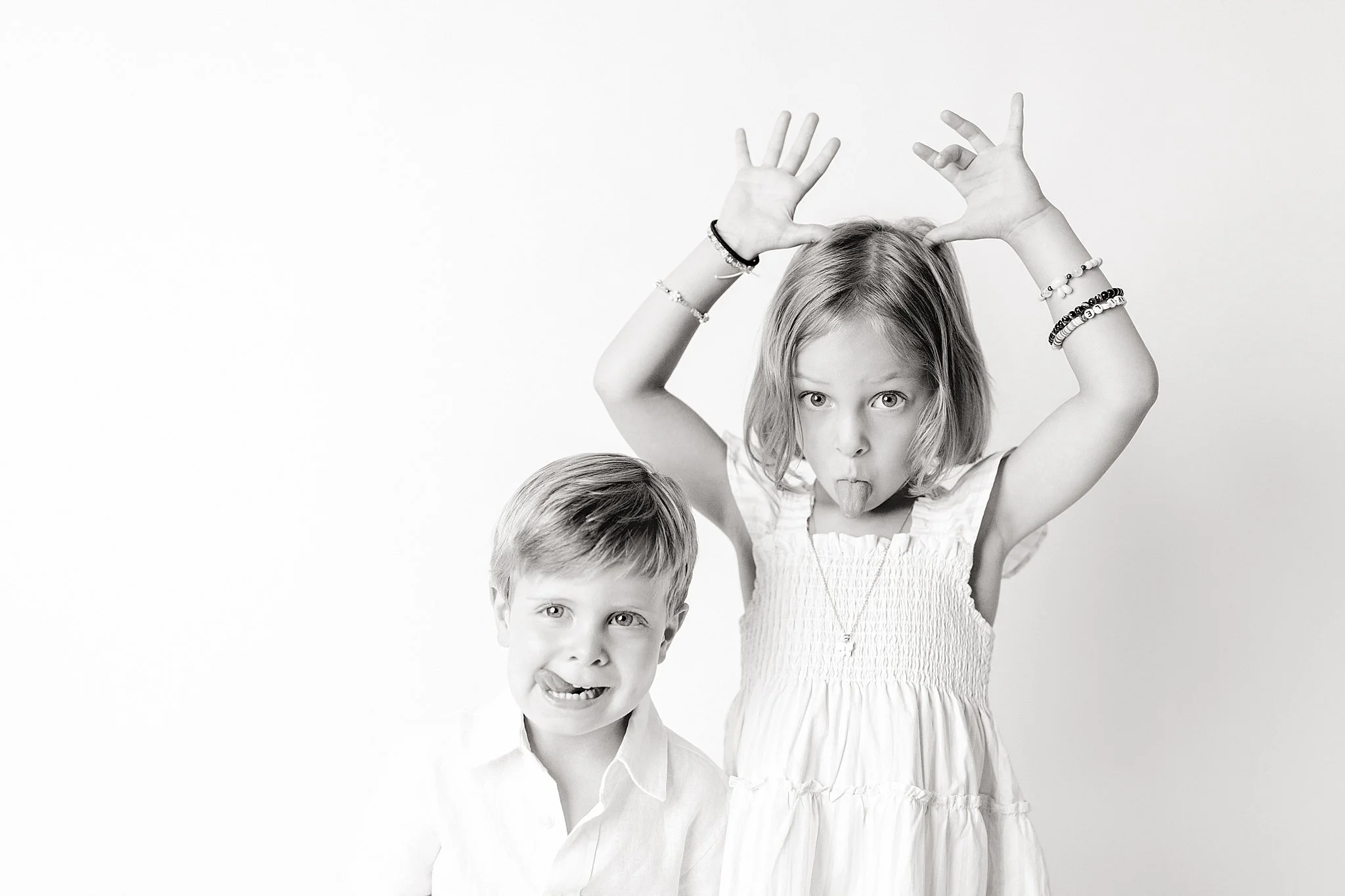 Black and white photo of two children, a girl and a boy. The girl is making a playful face with her tongue out and hands raised above her head with fingers spread, wearing bracelets and a dress. The boy is smiling, wearing a collared shirt, and posit