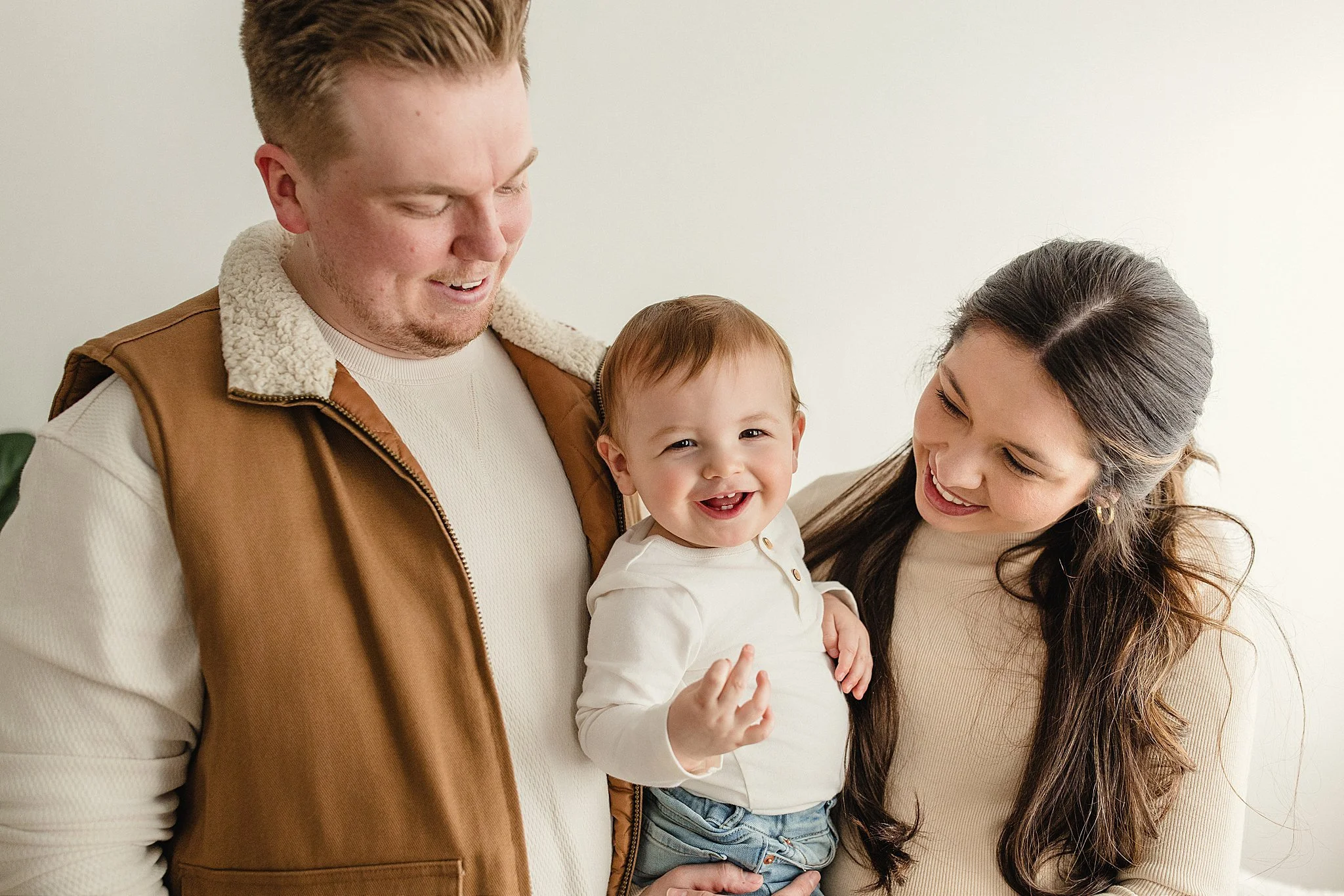 A happy family of three, with a man, woman, and a child, smiling and looking at each other indoors with a lifestyle photographer near me.