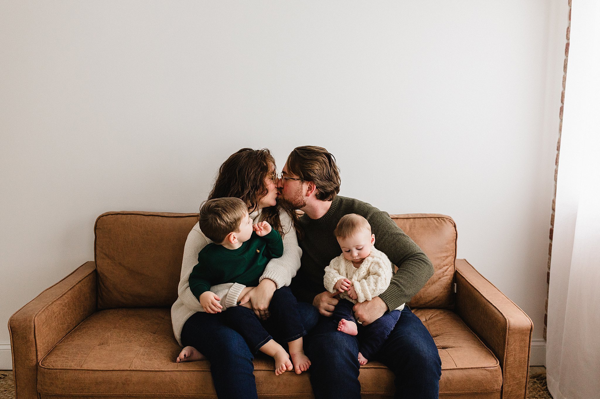 A family of four sitting on a brown sofa, with the parents kissing and the children sitting on their laps during a Naperville studio family photo session.