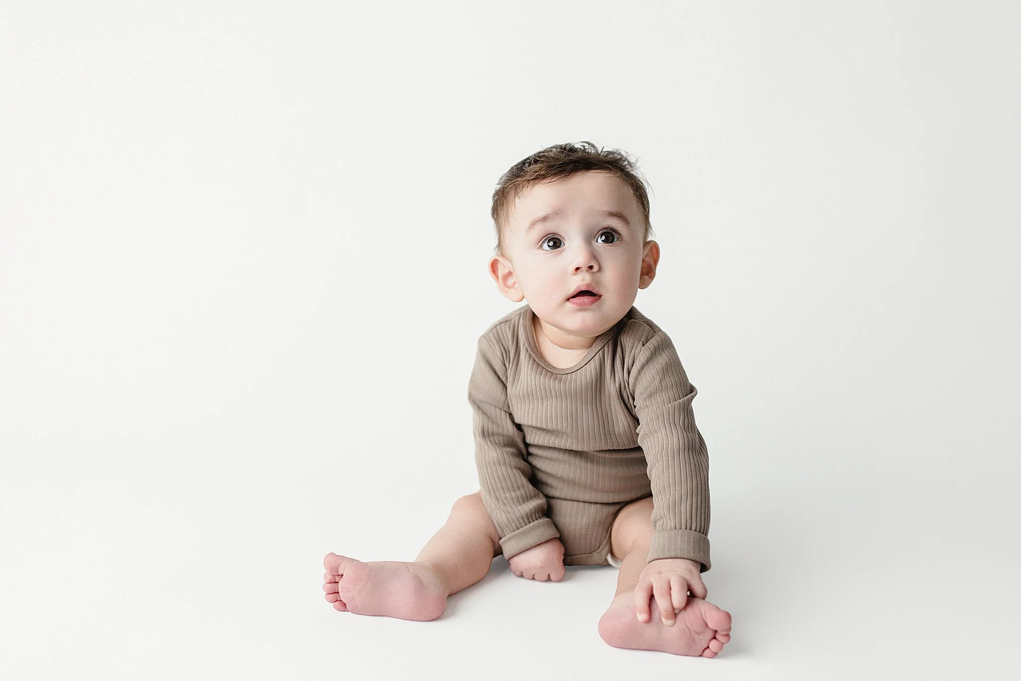 A young child with brown hair and big eyes sitting on a white surface, wearing a beige long-sleeve shirt and matching shorts, looking up with a curious expression during an infant photoshoot in Naperville, IL.