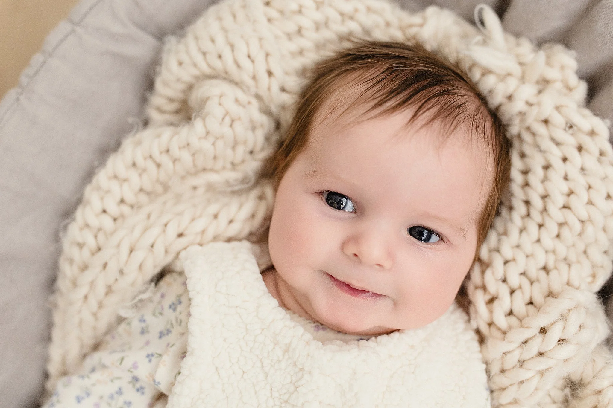 Close-up of a smiling baby with brown hair and blue eyes, wrapped in a chunky cream knit blanket and wearing a cozy cream bib, lying on a soft gray surface during newborn photography.