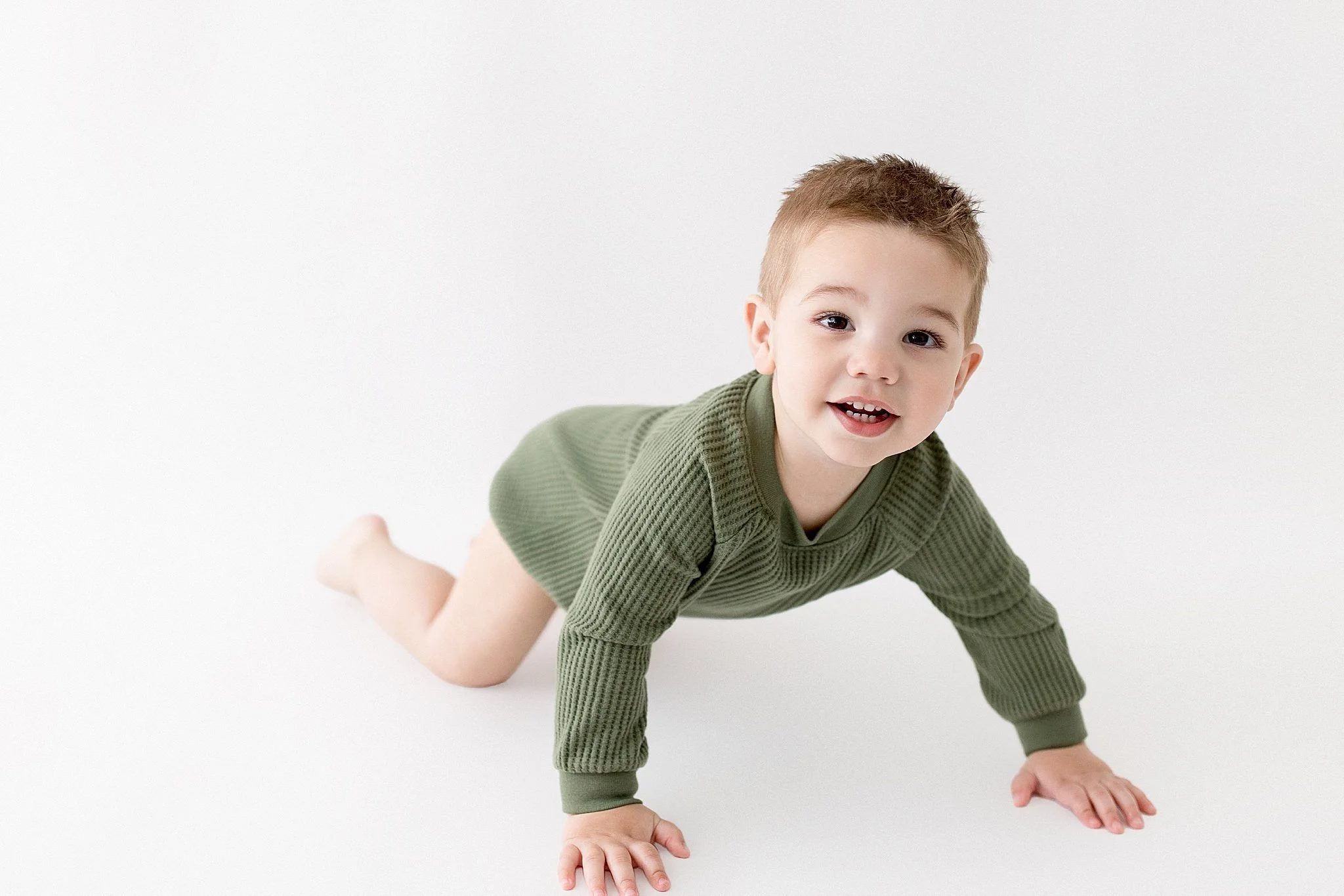 Young boy crawling on a white background, wearing a green long-sleeve shirt and smiling during baby photoshoot in downtown naperville, il.
