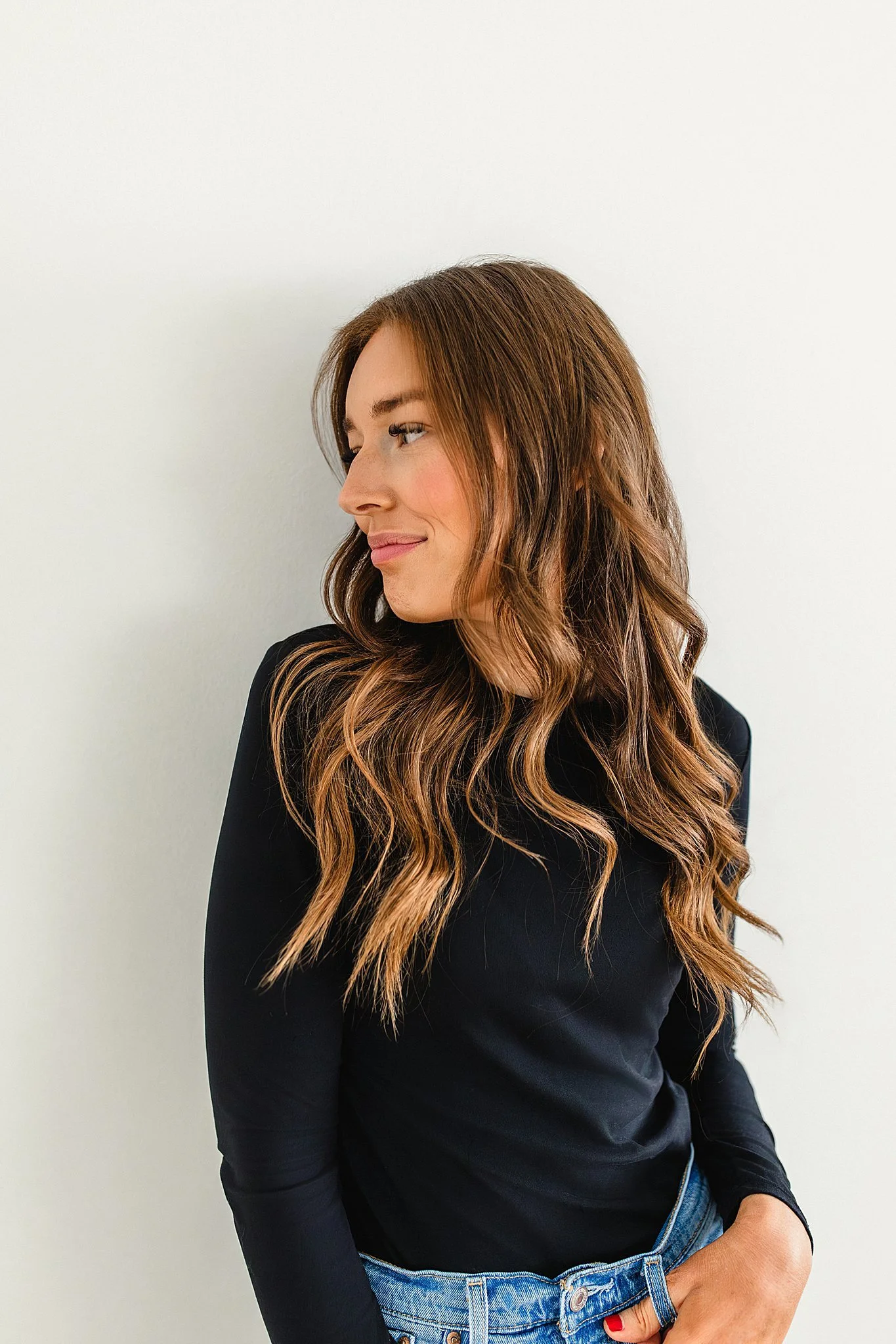 A young woman with long, wavy brown hair, wearing a black long-sleeve shirt and jeans, standing against a plain white wall, looking to her left with a slight smile during a brand photoshoot in Naperville, IL.