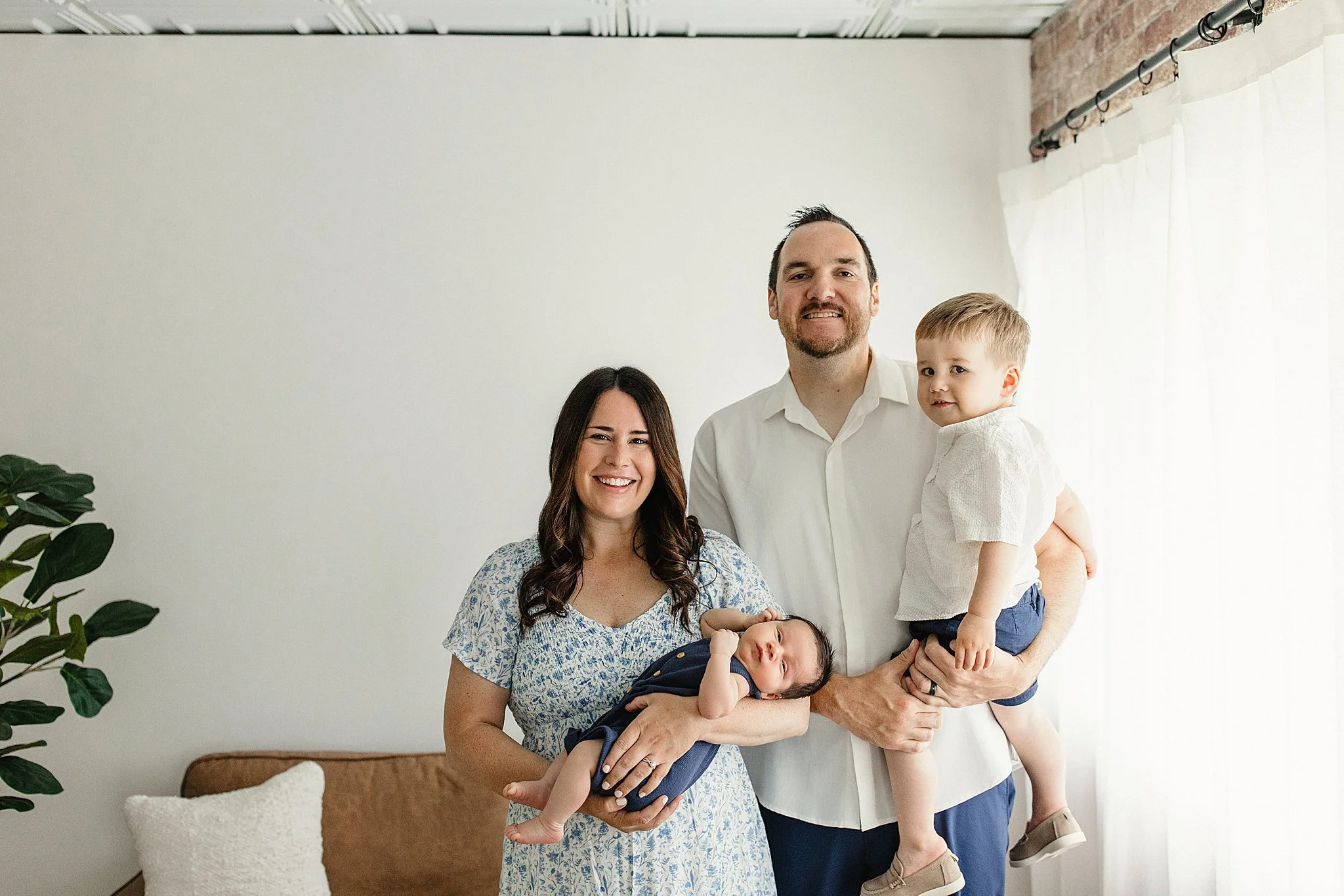 A family of four standing indoors in front of a white wall, with a woman holding a baby, a man holding a young boy, and a woman smiling at the camera with Marie Lopez photographer in Naperville, IL.
