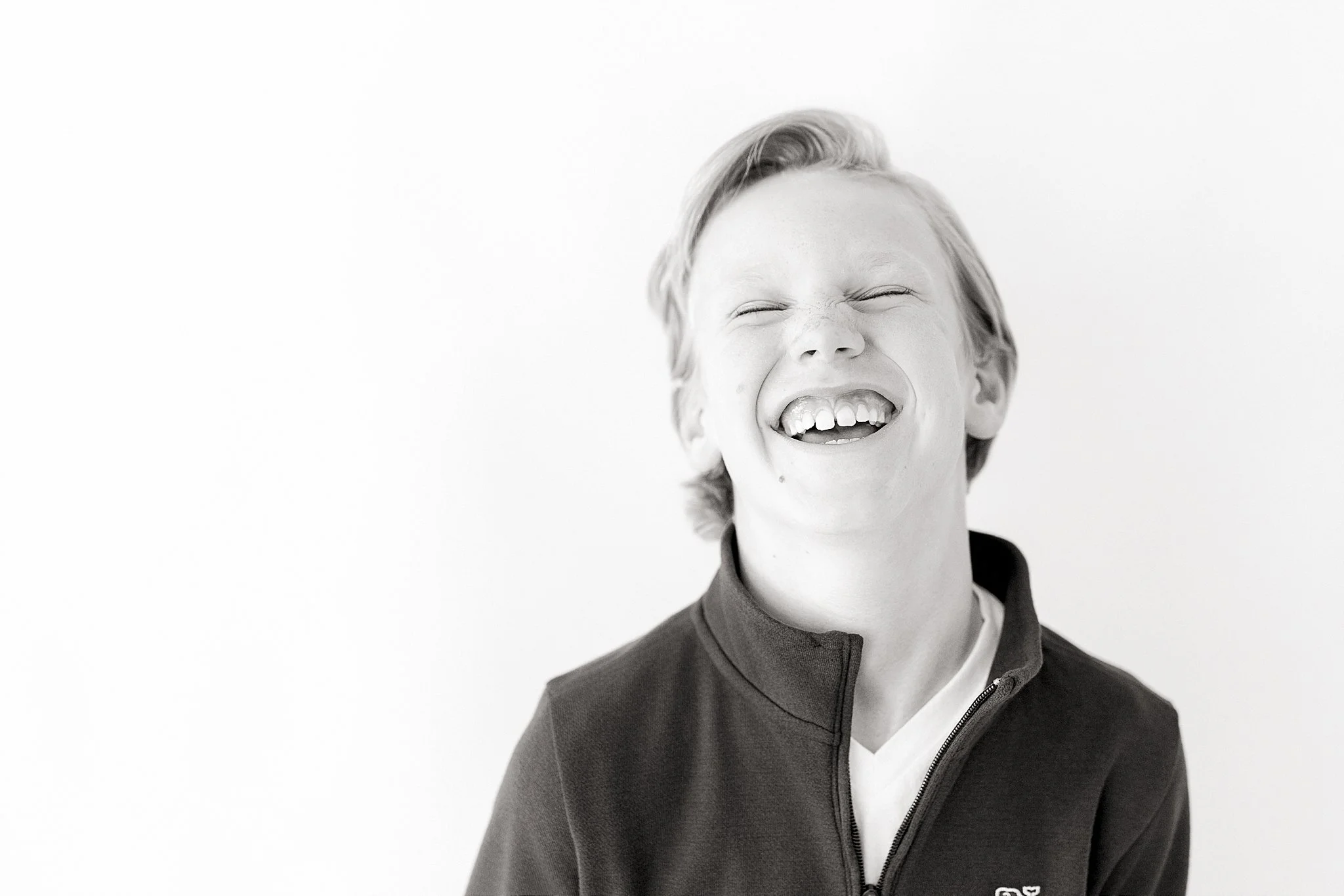Black and white photo of a young boy smiling widely with eyes closed, wearing a zip-up jacket and a white shirt inside, against a plain light background during personality minis naperville.
