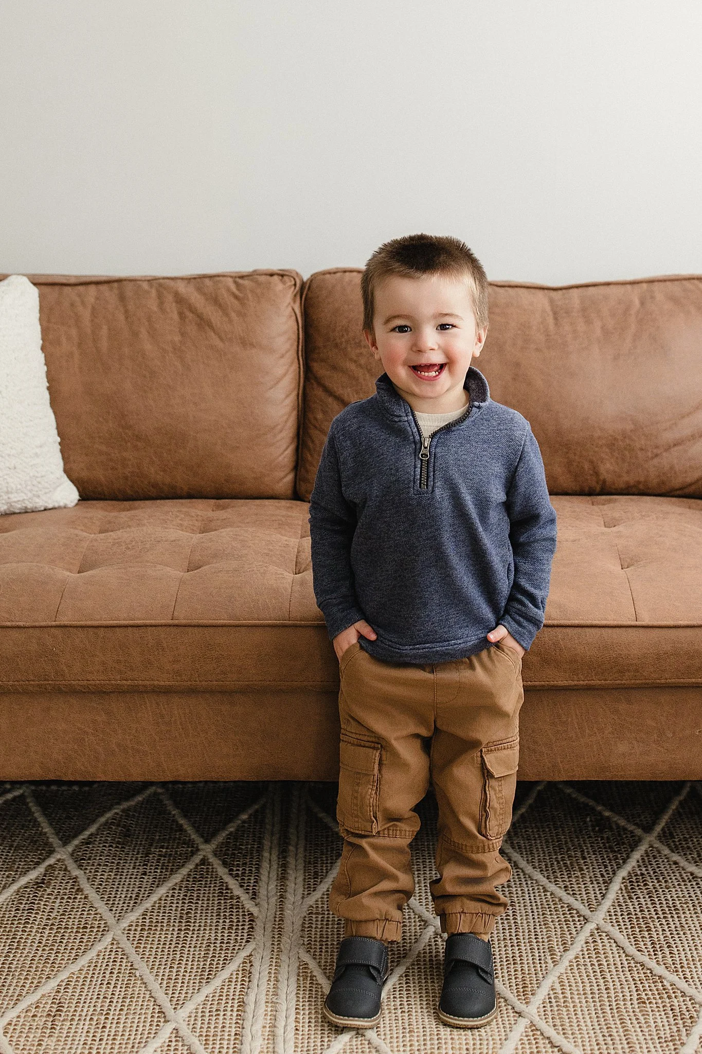 A young boy with short brown hair, wearing a navy blue zip-up sweater, tan cargo pants, and black shoes, standing in front of a brown leather sofa inside a home with a naperville photographer.