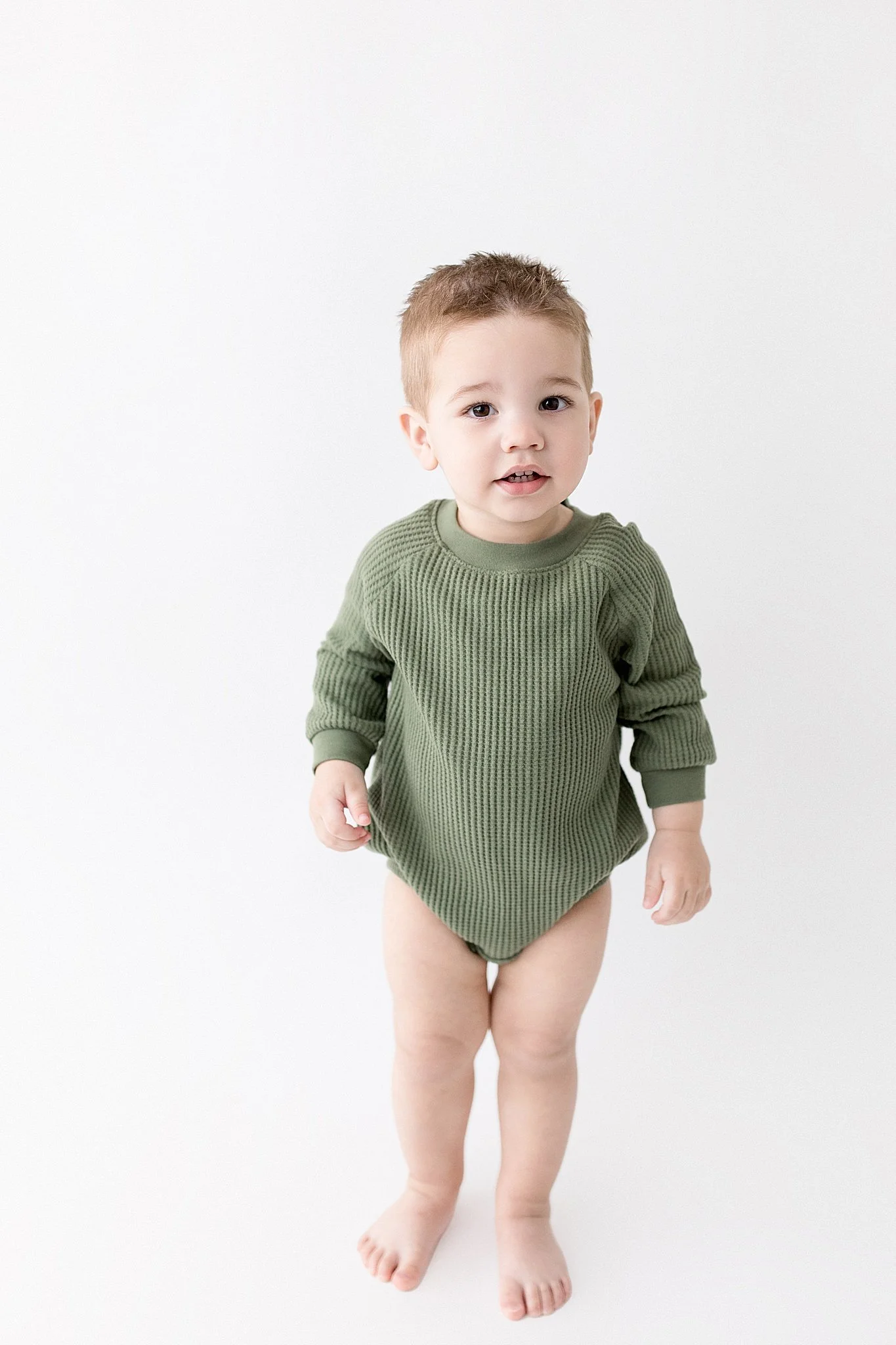 A young toddler with short brown hair, wearing a green textured long-sleeve shirt, standing barefoot on a white background, looking at the camera with the best baby photographer in Naperville, IL.