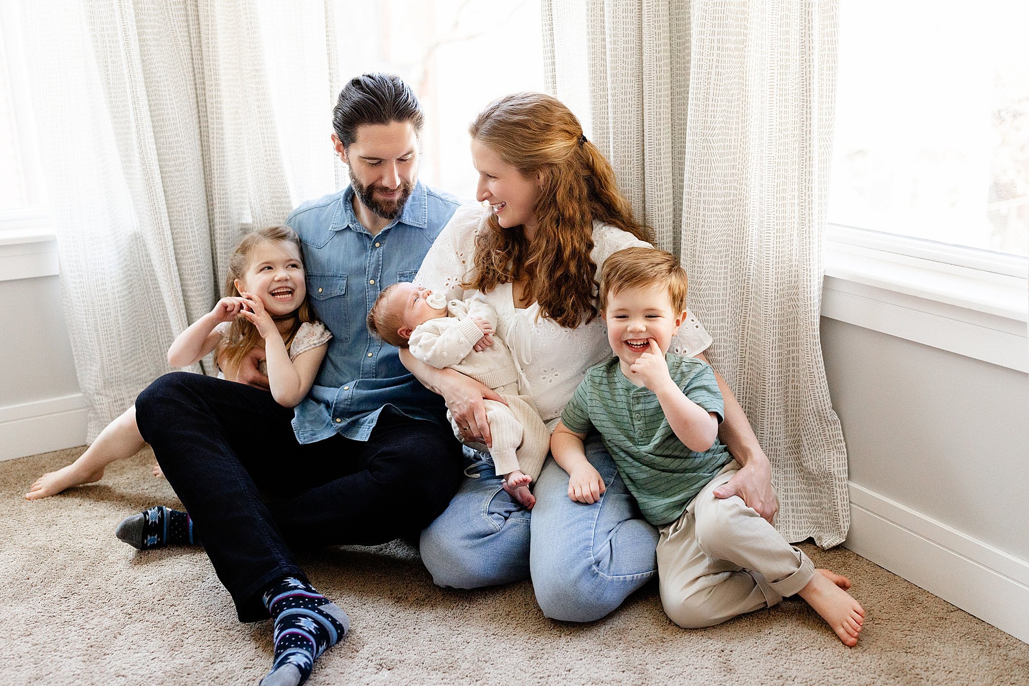 A family of five, including a mother, father, two sons, and a baby, sitting on the carpet in a room with light-colored curtains and windows, smiling and enjoying time together.
