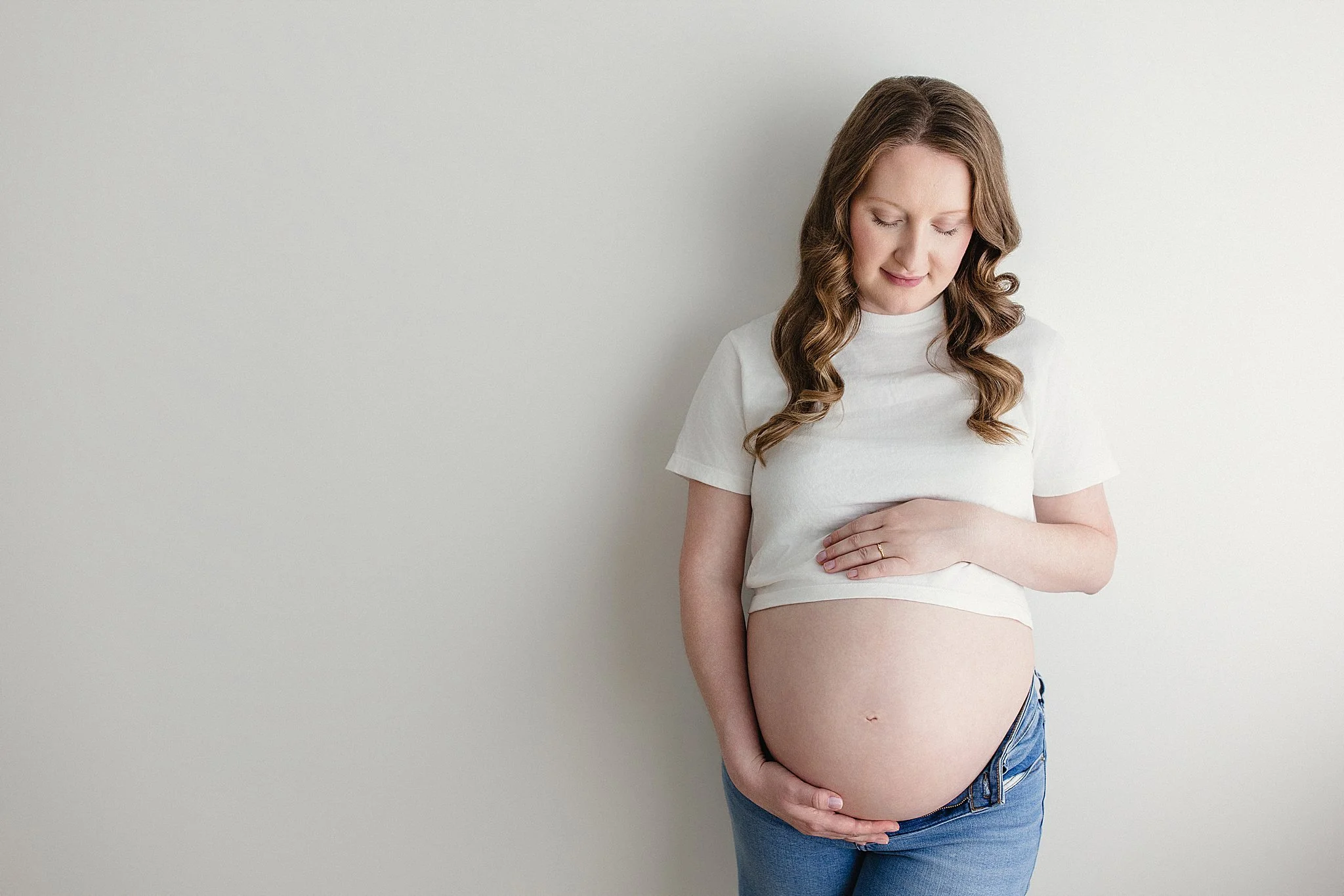 A pregnant woman standing against a plain white wall, wearing a white t-shirt and blue jeans, with her eyes closed and hands resting on her belly with Naperville maternity photographers.