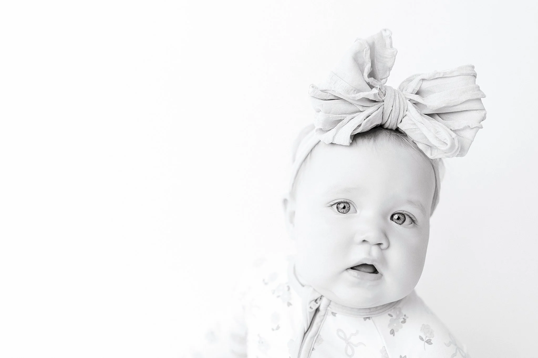 A young child with light-colored eyes, partially visible face, wearing a light-colored jacket with a floral pattern and a large, fabric bow tied around the head in a headband on a white background with the best baby photographer in Naperville, IL.