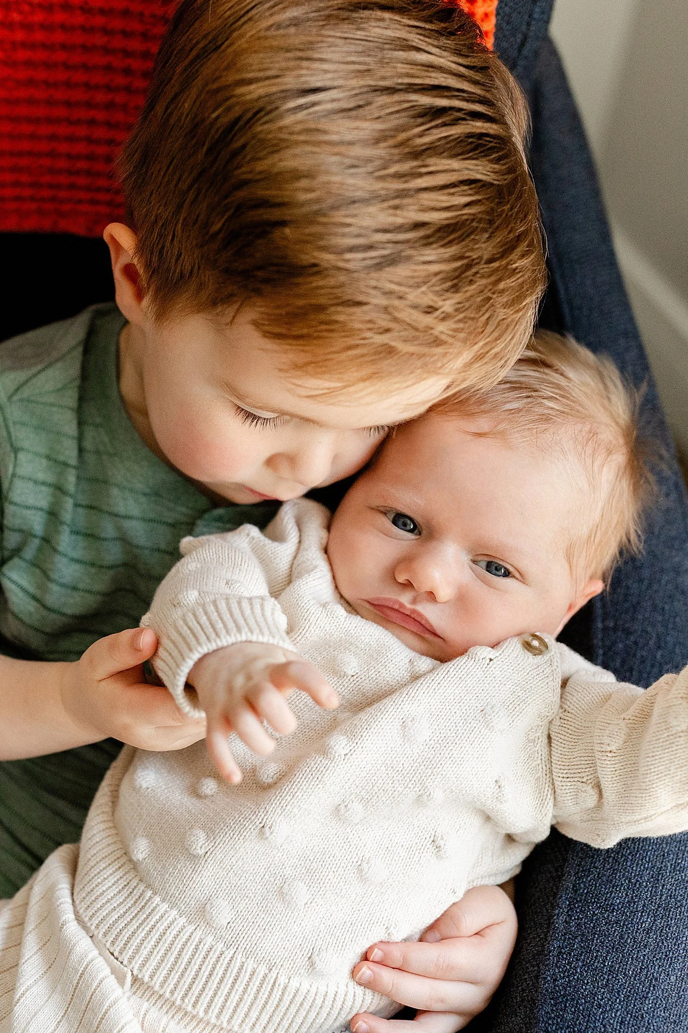 A young boy with light brown hair and fair skin hugging a baby with blonde hair and fair skin, both lying on a person's lap. The boy is wearing a green striped shirt and the baby a cream-colored sweater.