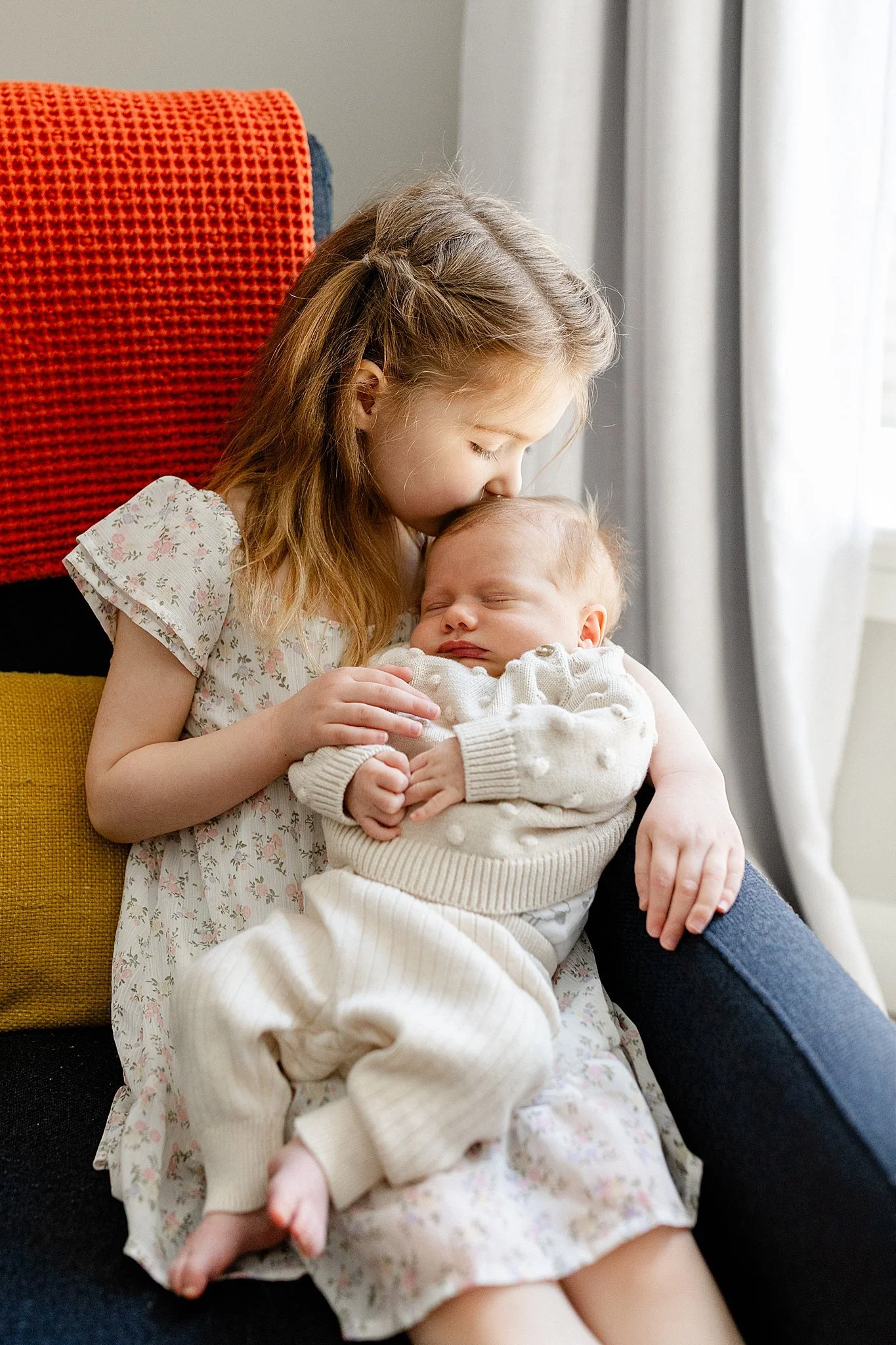 A young girl with long, light brown hair kissing a sleeping baby with a reddish-blonde hair on the top of their head, sitting on a yellow cushion in a room with a window and white curtains with Ally and B Photography.