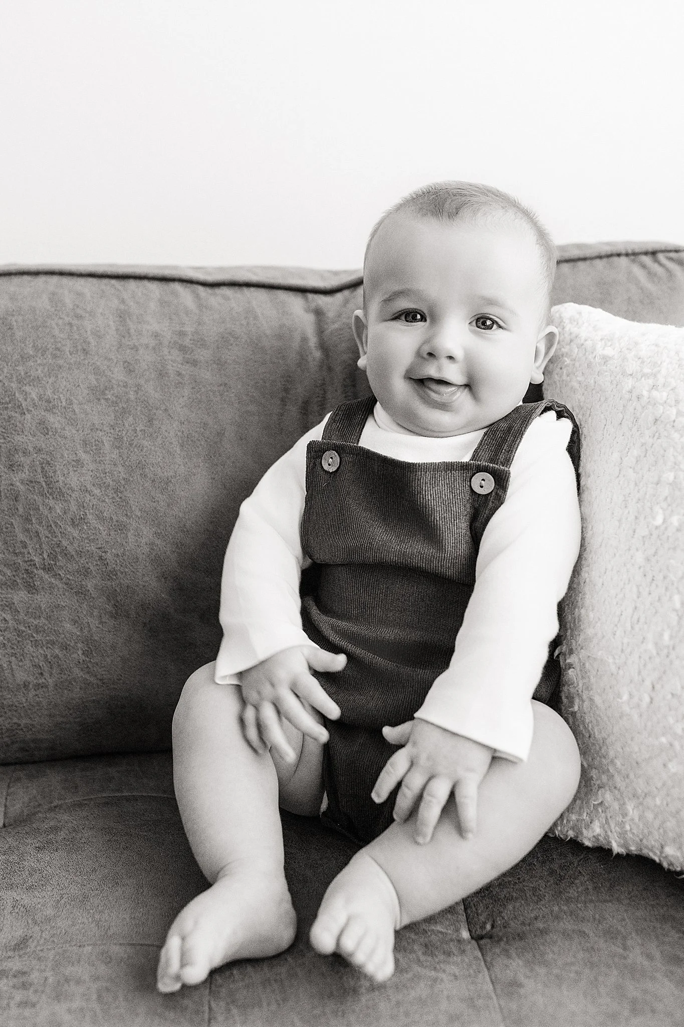Black and white photo of a smiling baby sitting on a couch, wearing a long-sleeve shirt and overalls with best baby photographer in Naperville, IL.