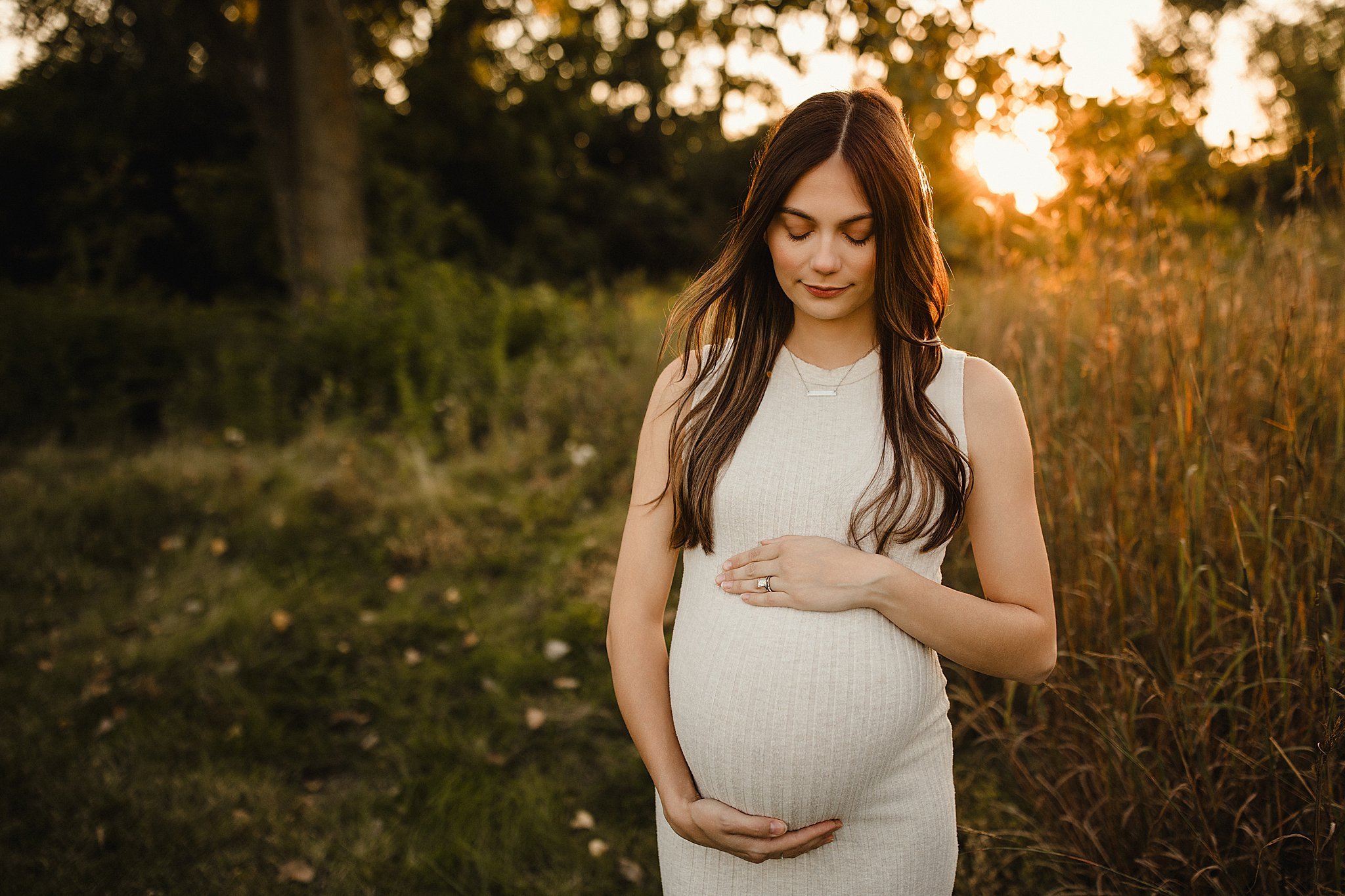 A pregnant woman standing outdoors during sunset, gently holding her belly with both hands, wearing a sleeveless beige dress, with a peaceful expression and eyes closed during a golden hour maternity photo session in Lisle, IL.