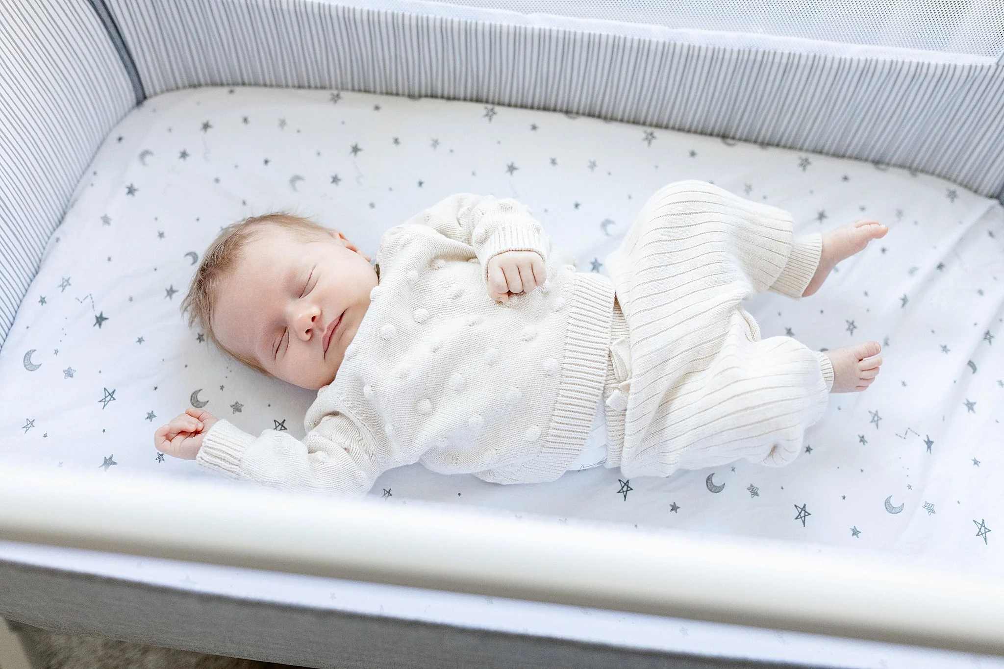 A sleeping baby girl in a white sweater and pants lying in a crib with star and moon patterned bedding with newborn photographers Naperville.