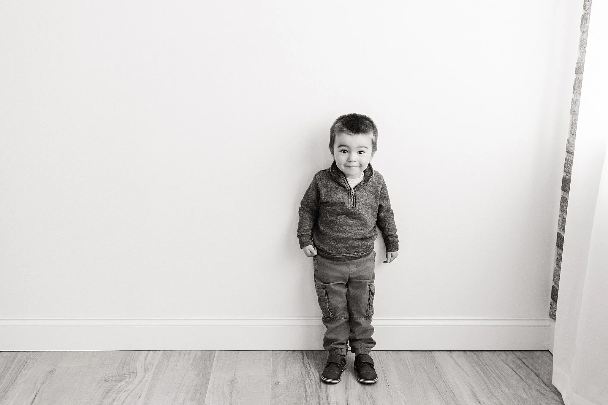 A young boy standing indoors against a plain wall, smiling and looking at the camera with Ally and B photography for a kids photo session.