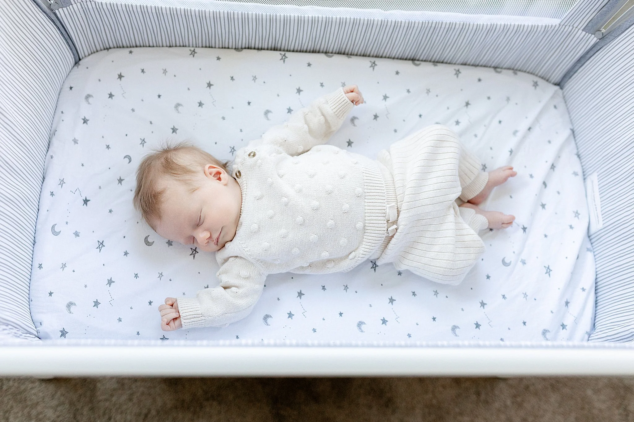 A sleeping baby wearing a white sweater and matching pants, lying on a star and moon patterned sheet inside a crib with striped sides.