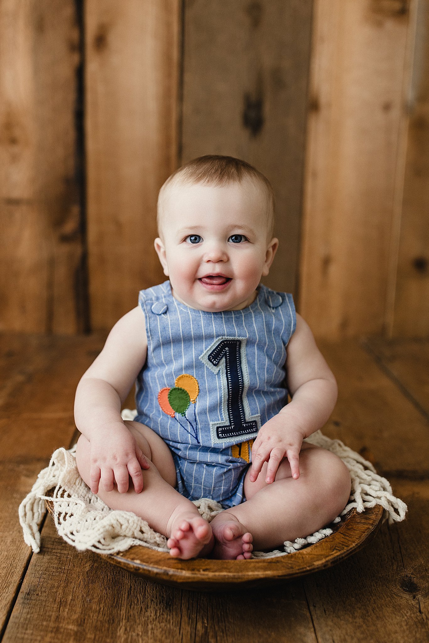 A smiling baby sitting on a wooden floor in front of a wooden background, wearing a blue striped outfit with the number 1 and balloons on it during a first birthday photoshoot with Ally and B Photography in downtown Naperville, IL.
